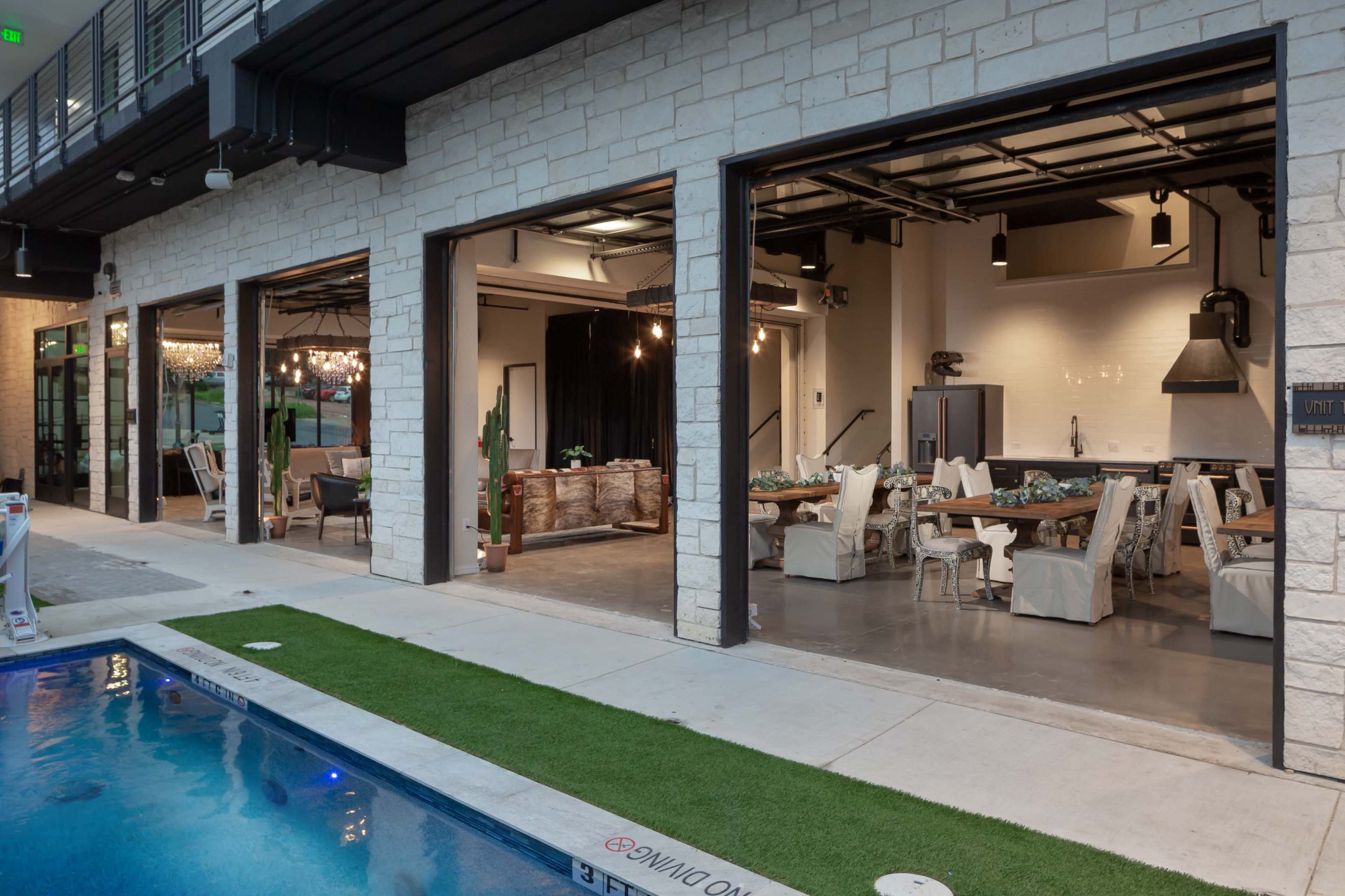 The image shows a modern indoor-outdoor space featuring a dining area with tables and chairs, adjacent to a swimming pool.