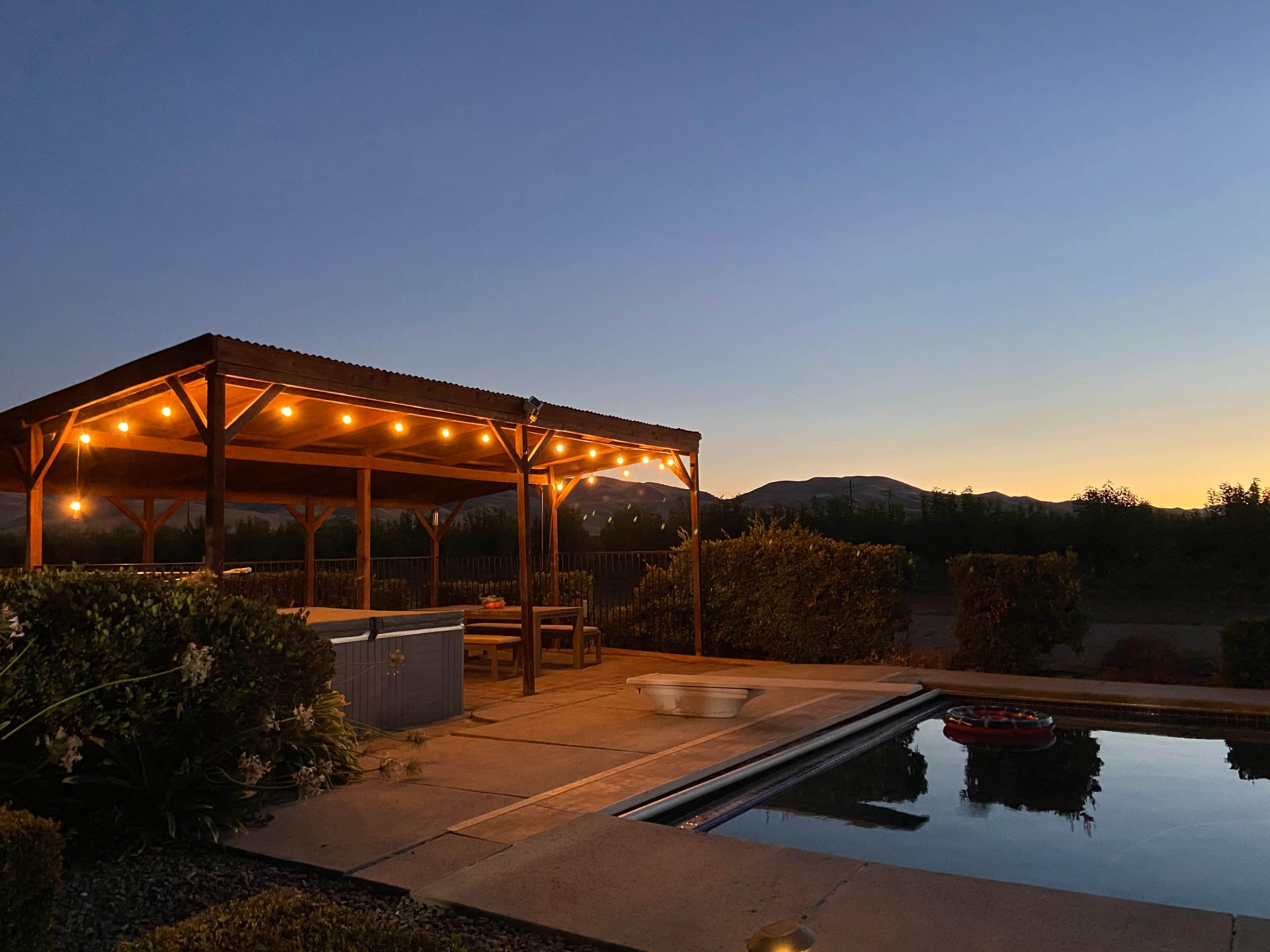 A wooden patio with string lights overlooks a pool and mountains at dusk.