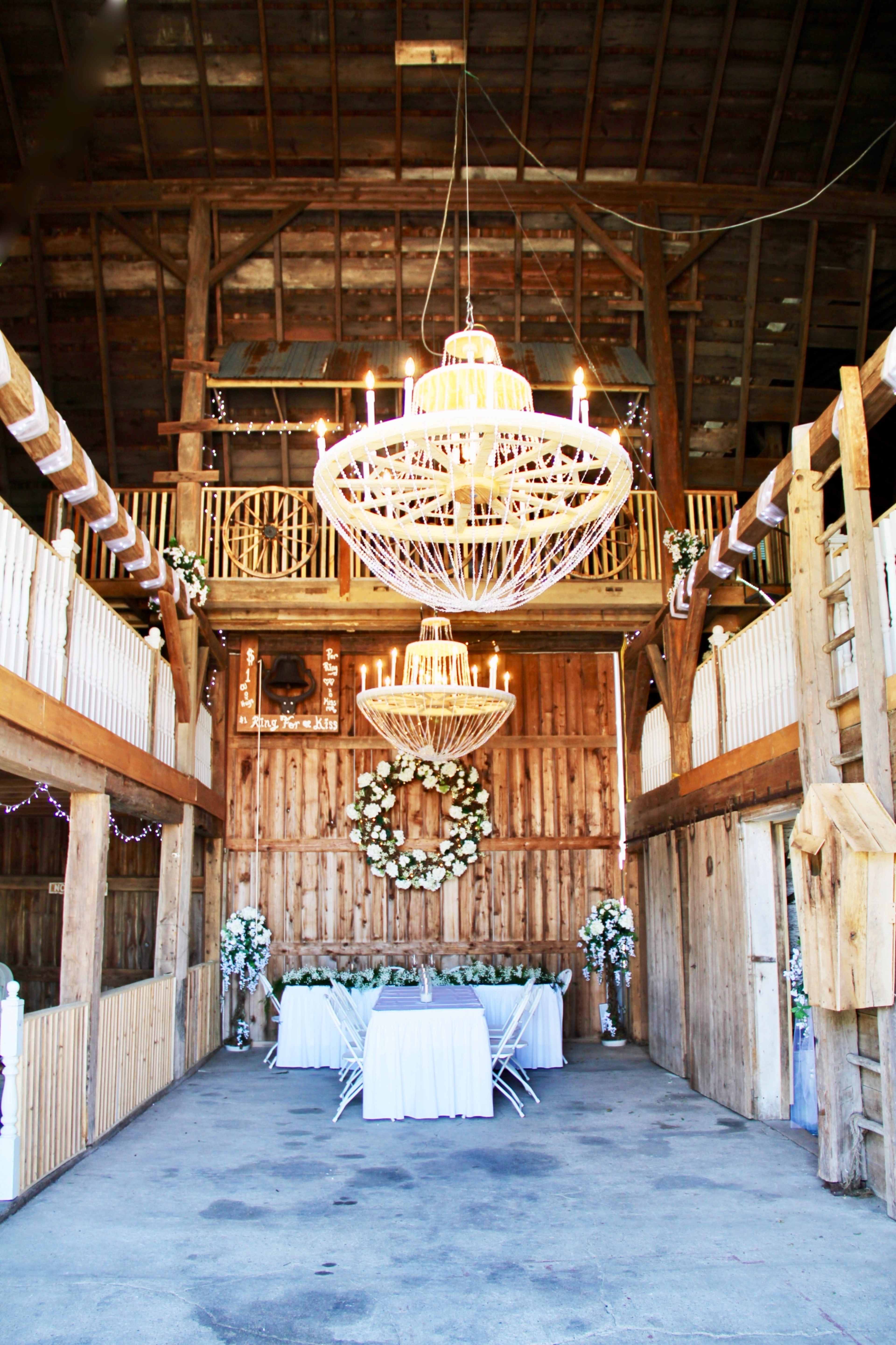 The interior of a barn features two large chandeliers, wooden walls, and a decorated table set for a wedding reception.