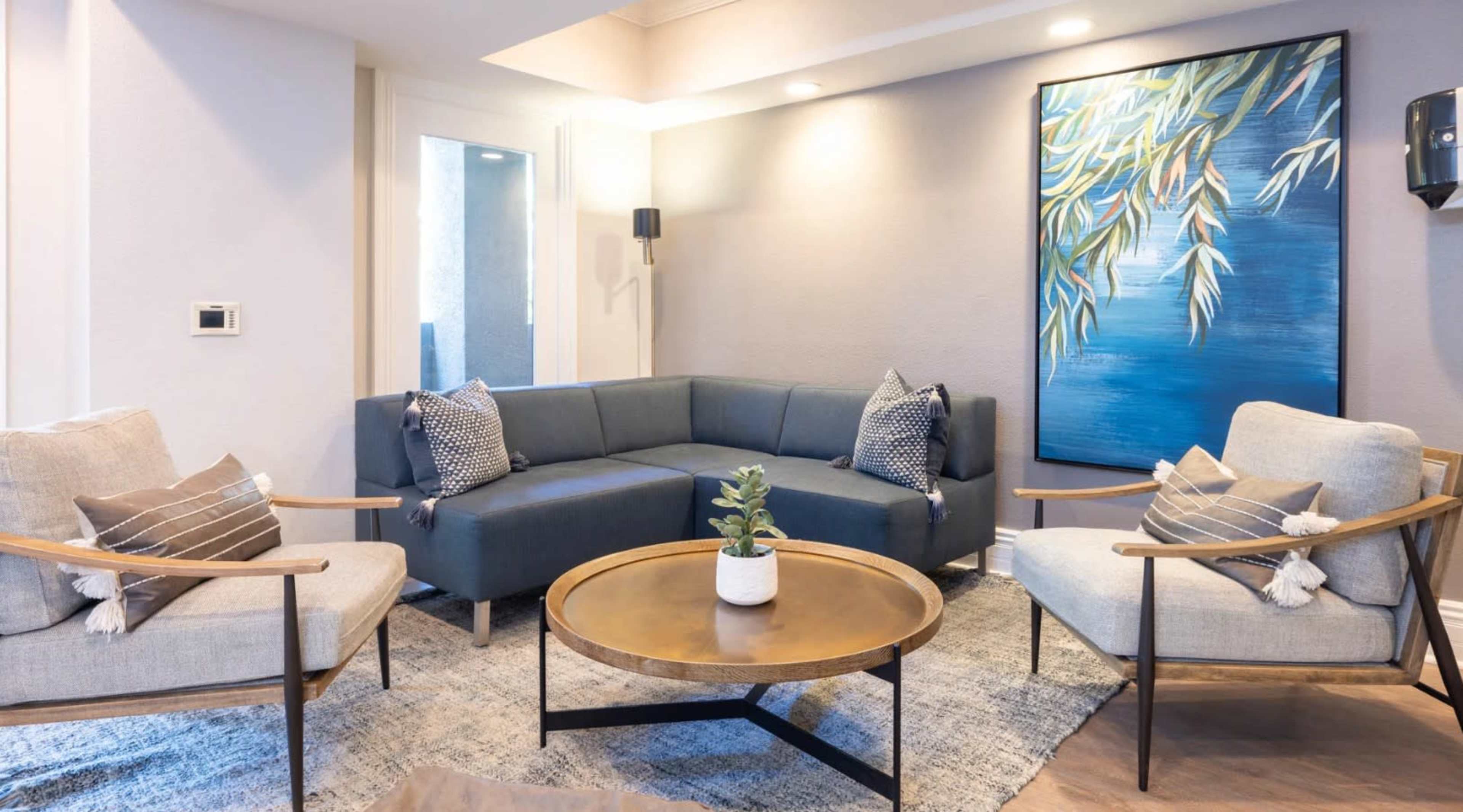The image shows a modern living room setup featuring a blue sectional sofa, two light-colored armchairs, a round coffee table, and a large painting with blue and green tones on the wall.