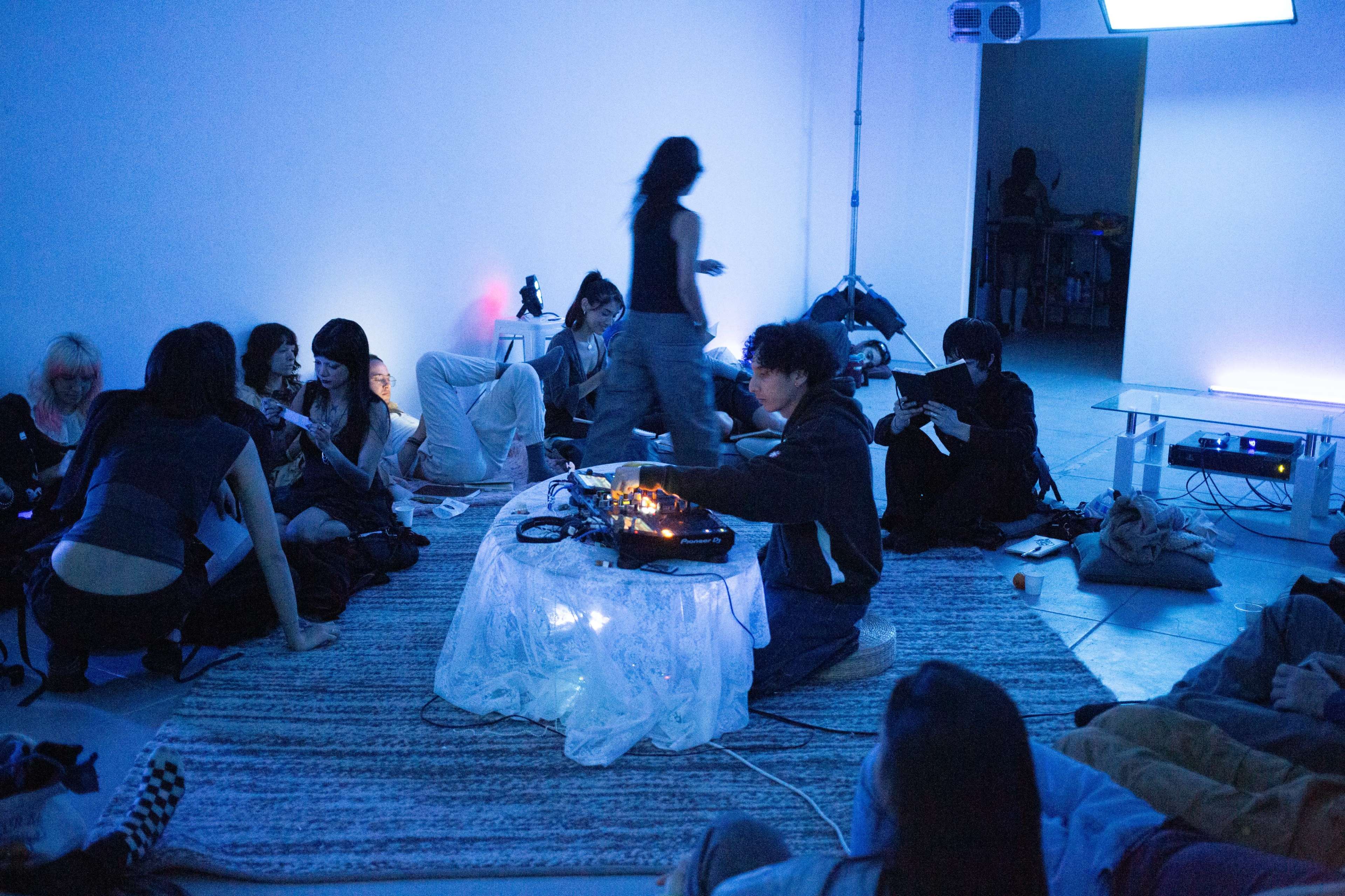 A group of people are sitting on the floor in a dimly lit space, with one person performing at a table while others socialize around them.