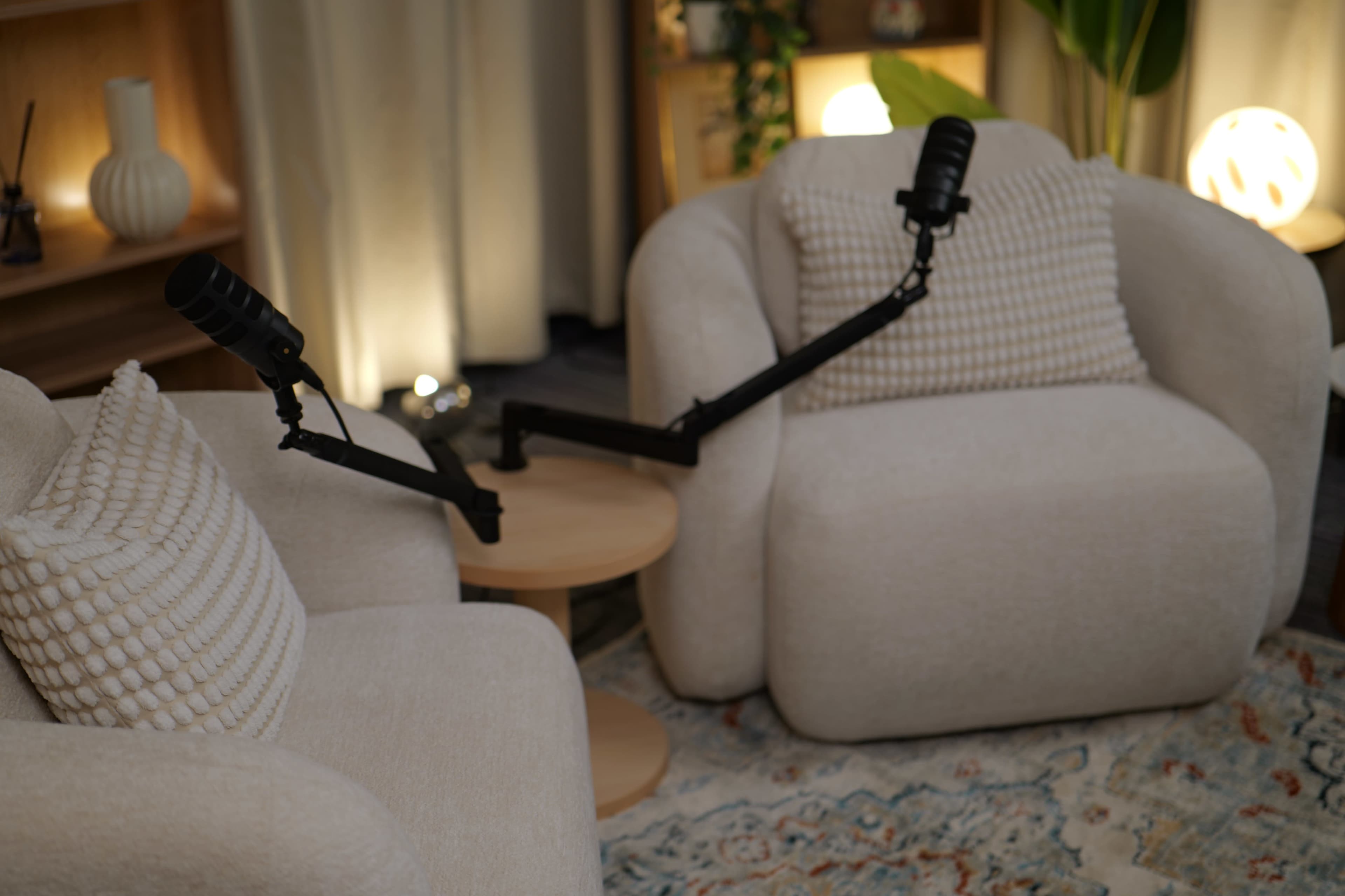 The image shows two cozy armchairs with microphones mounted on adjustable arms, positioned near a small wooden table in a warmly lit room.