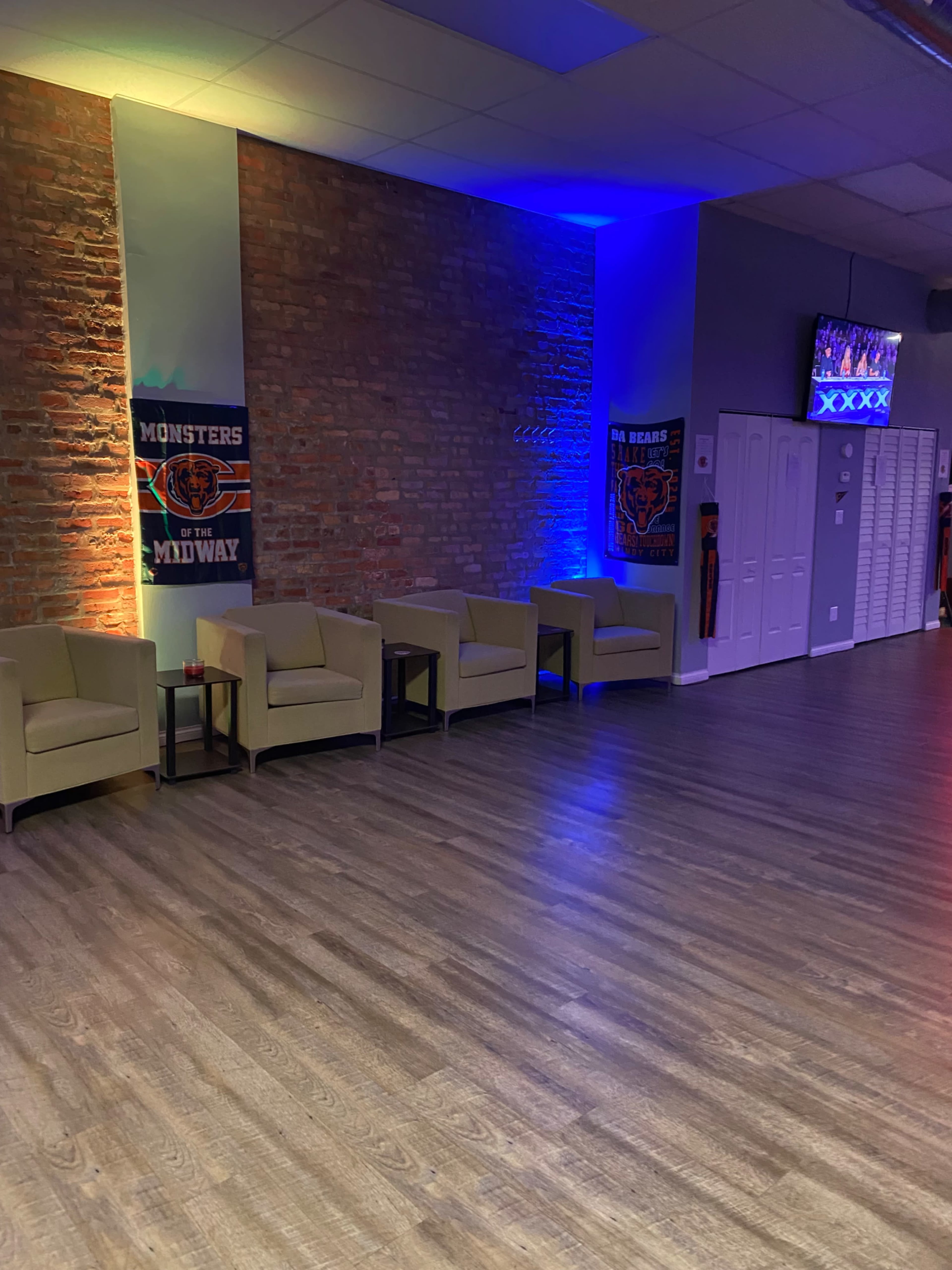 The image shows a spacious room with wooden floors, featuring a row of green chairs against a brick wall, illuminated by blue and purple lights, with team banners on display.