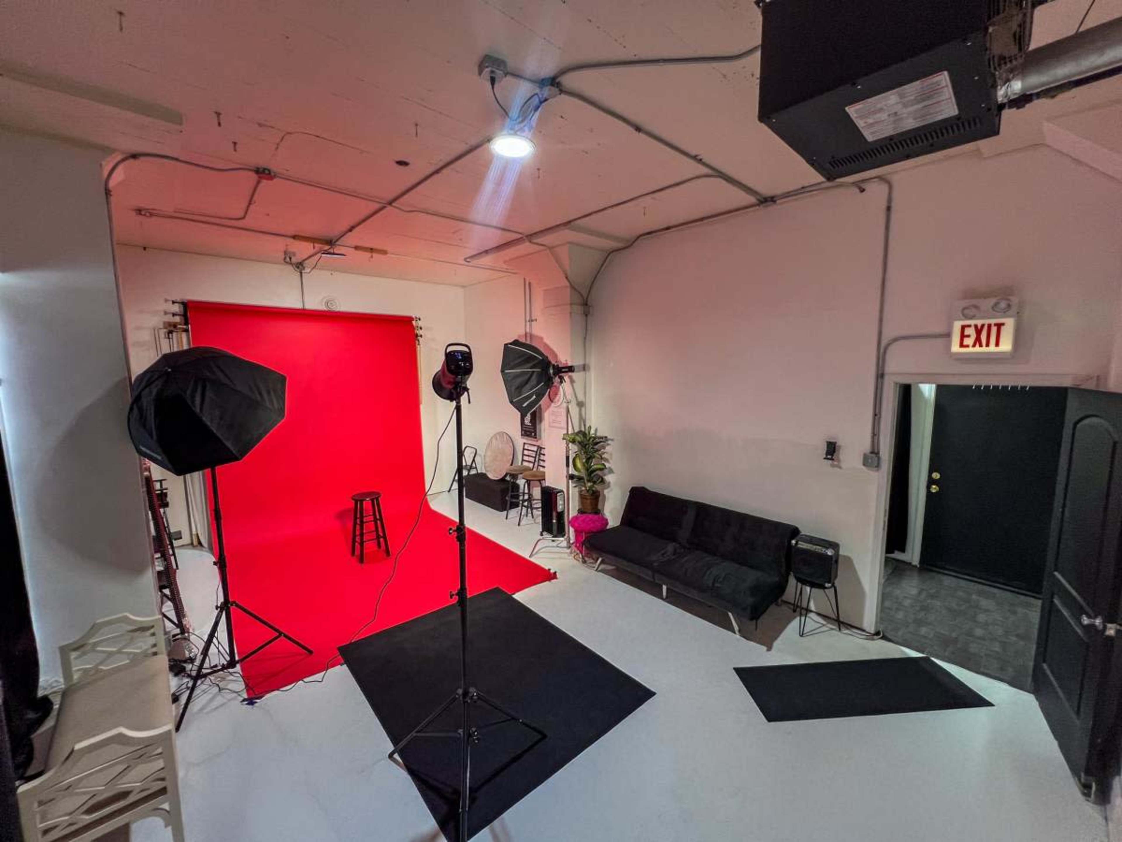 The image shows a photography studio with a red backdrop, lighting equipment, a black couch, and a stool positioned on a white floor.