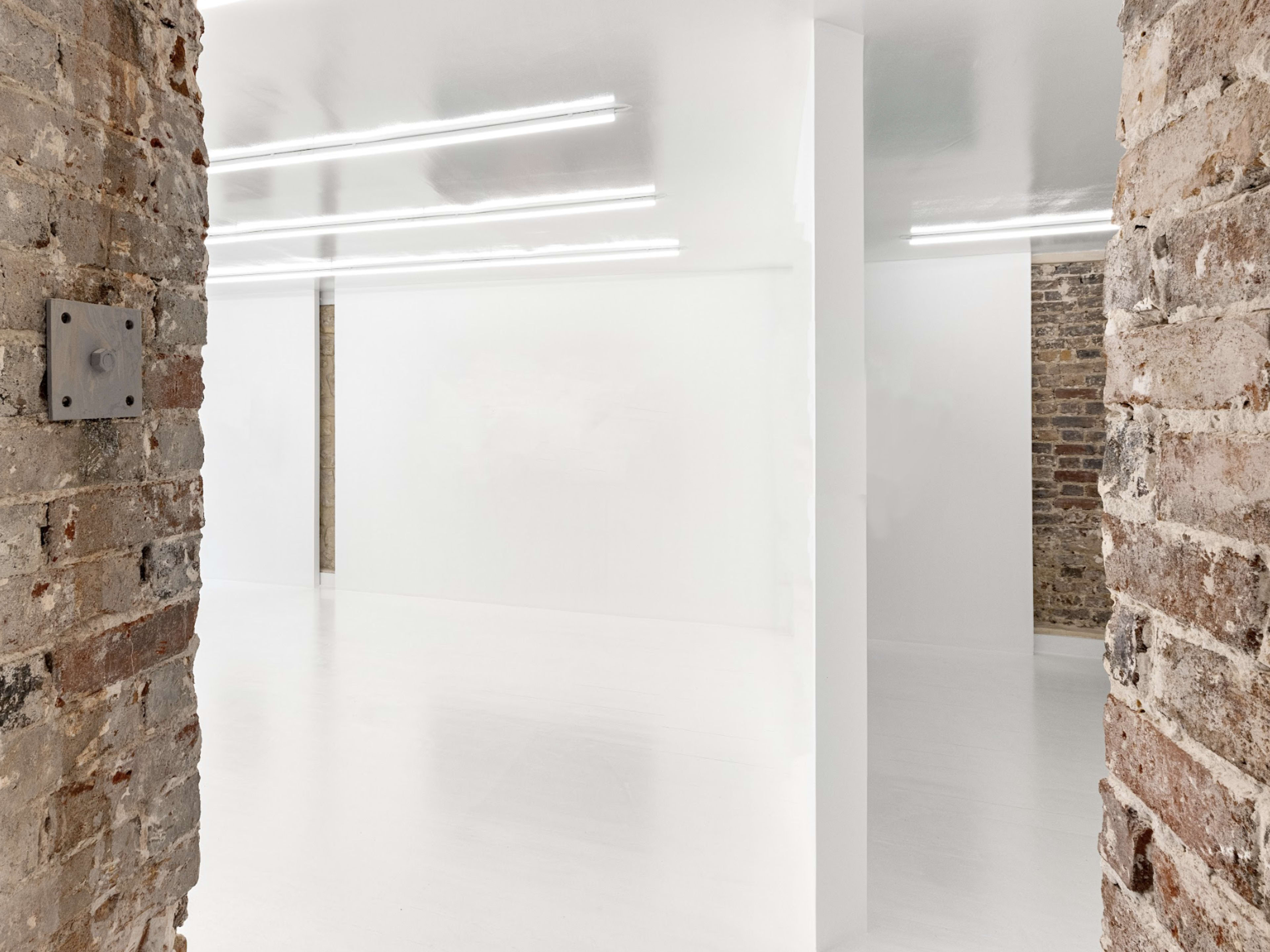 The image shows a spacious, minimalist interior with white walls and a glossy floor, framed by exposed brick columns.