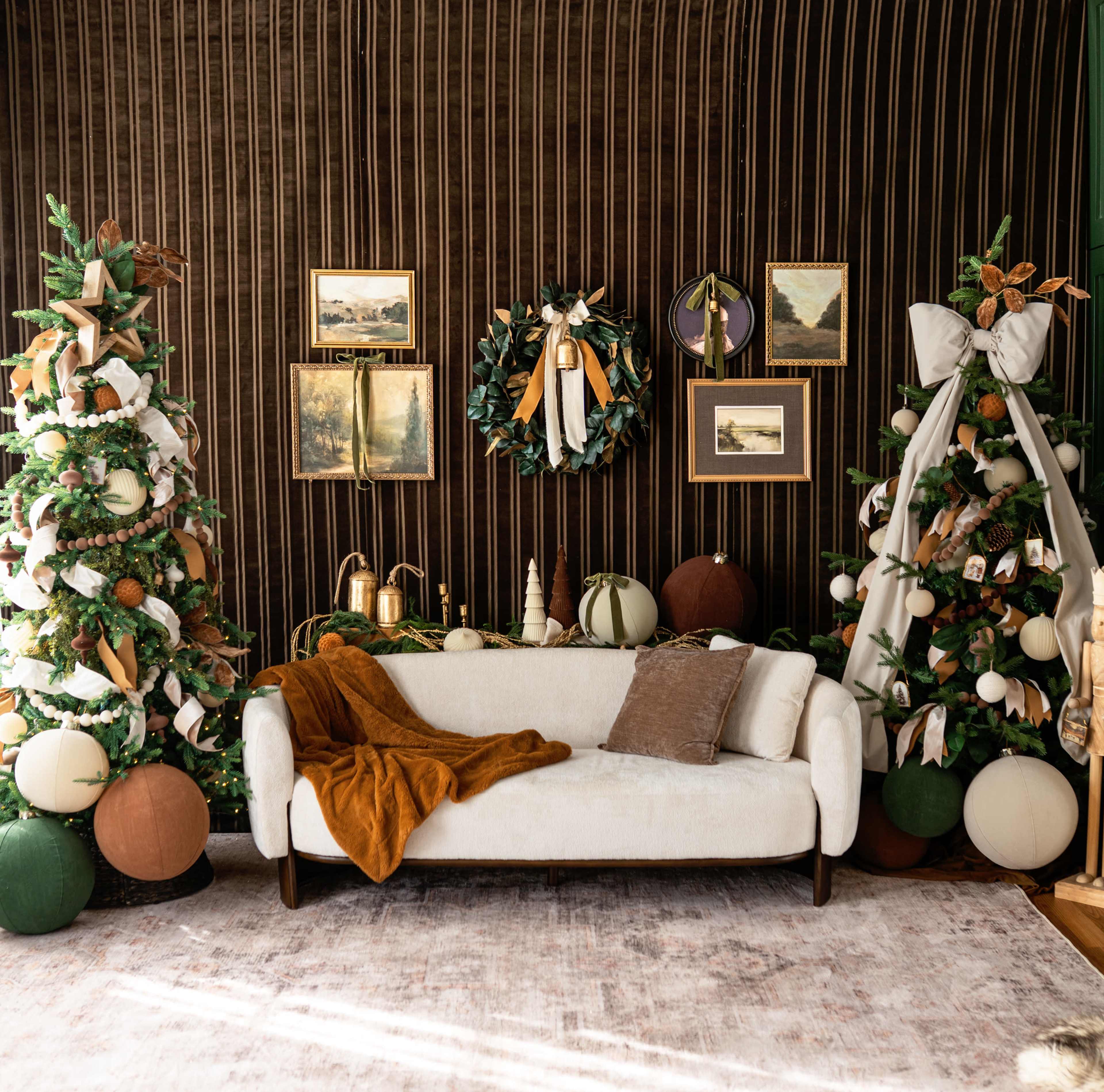 A cozy living room is decorated for the holidays, featuring a cream-colored sofa surrounded by festive garlands, decorated trees, and framed artwork on a wooden wall.
