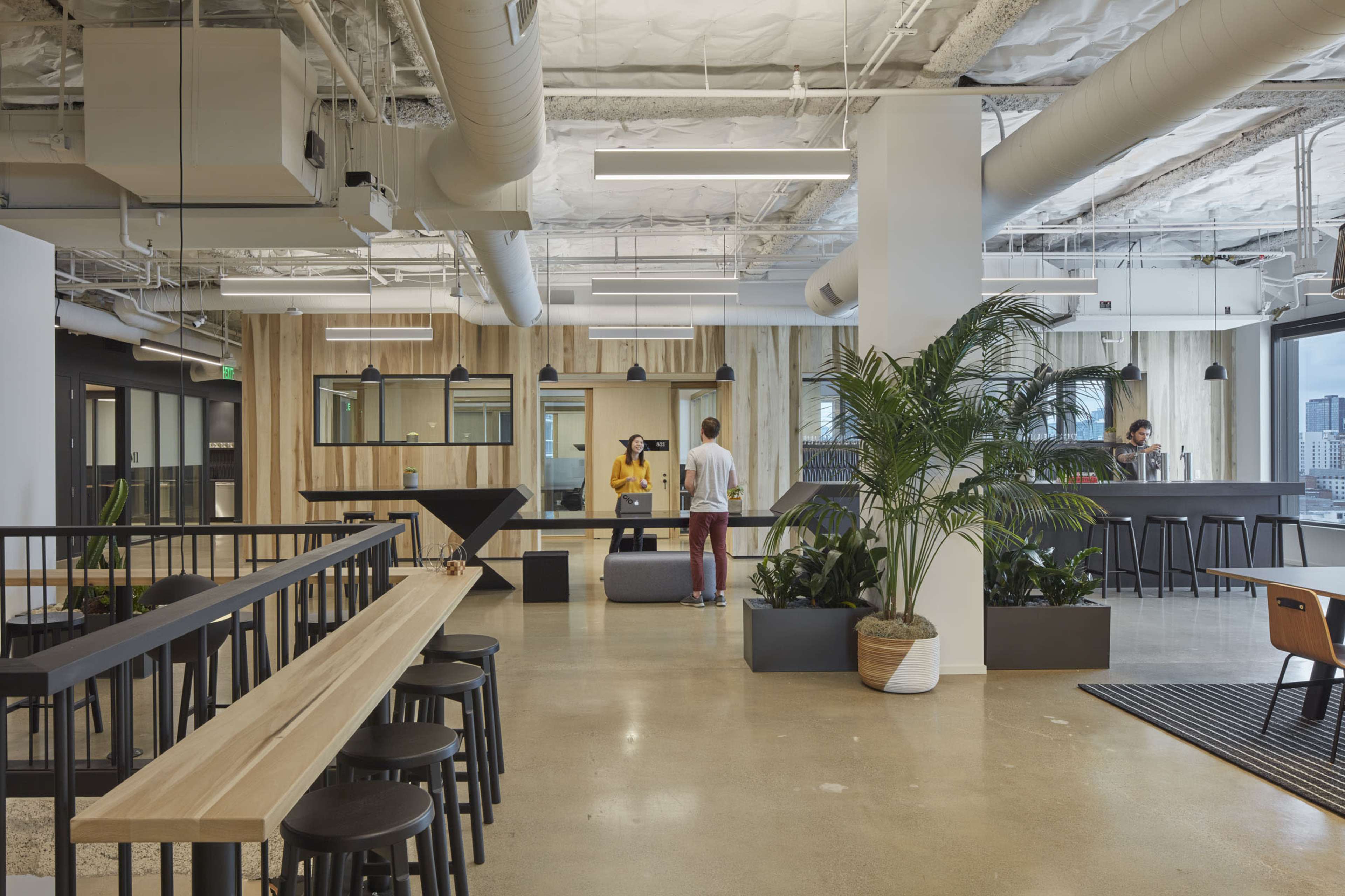 A modern office space featuring a large wooden table, plants, and employees engaged in various activities, with minimalistic design elements and open areas.