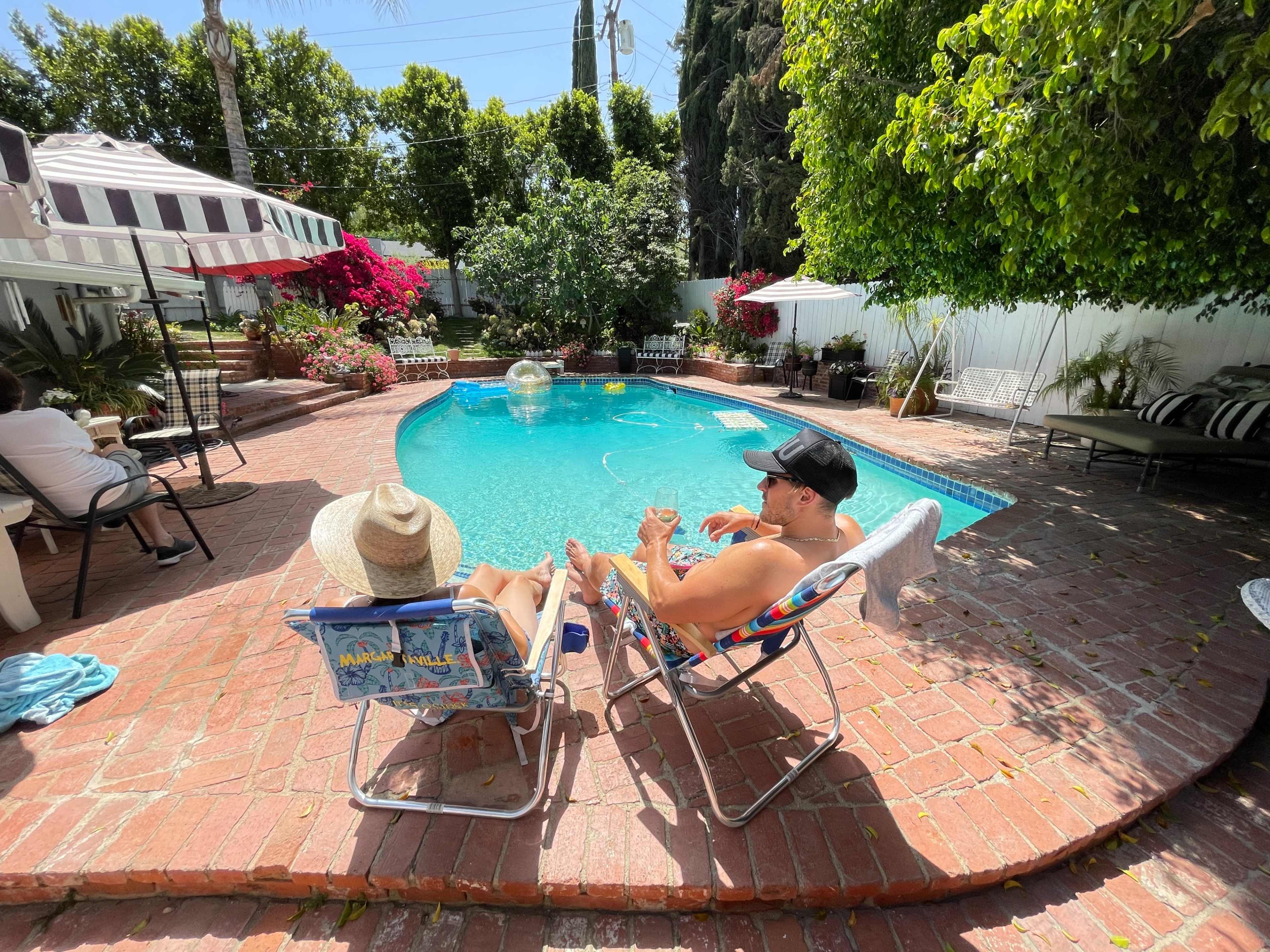 Two people relax in lawn chairs by a swimming pool surrounded by tropical plants and brick patio.