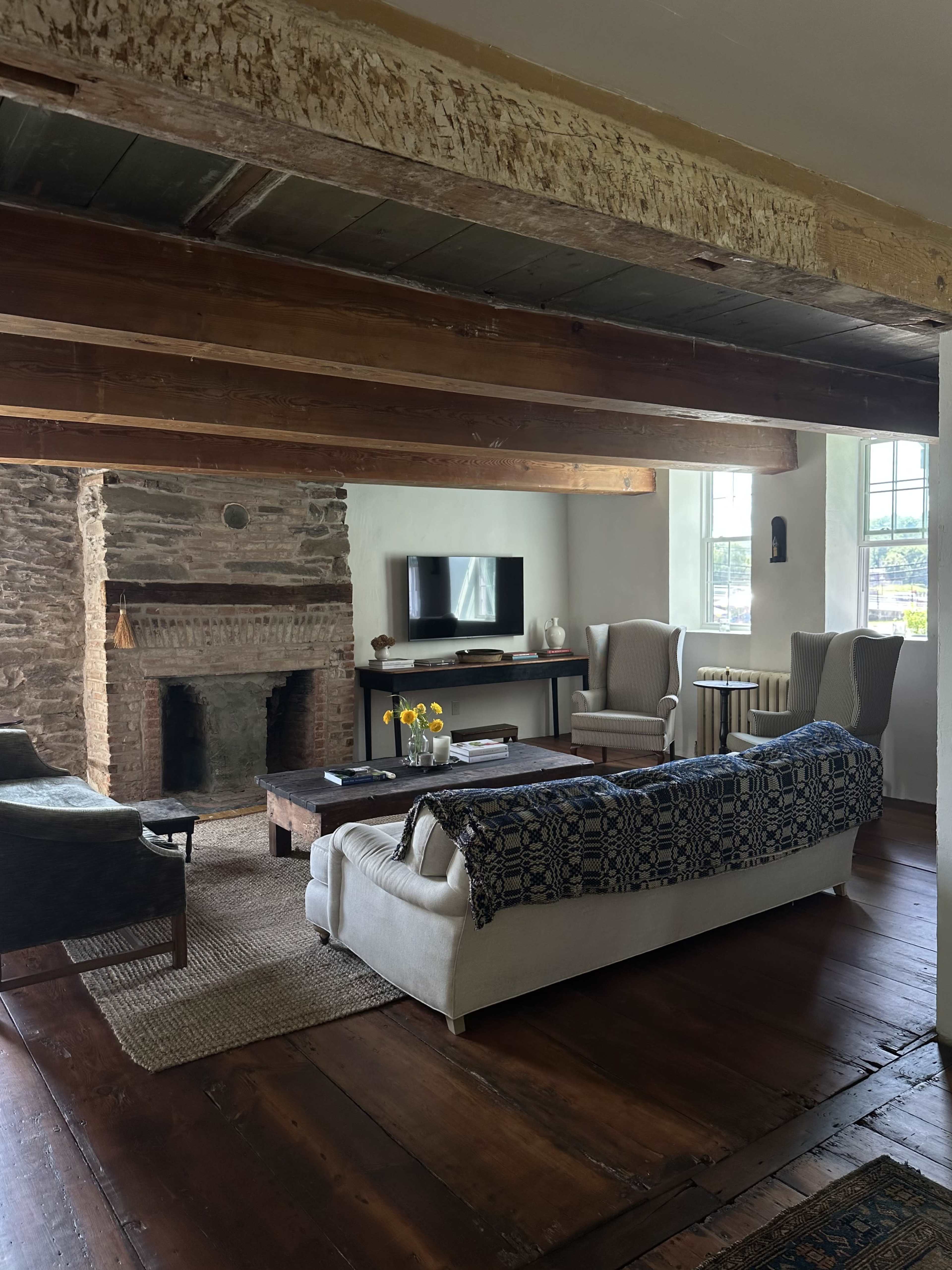 The image shows a cozy living room with wooden beams, a stone fireplace, a television, and two armchairs next to a coffee table adorned with fresh flowers.