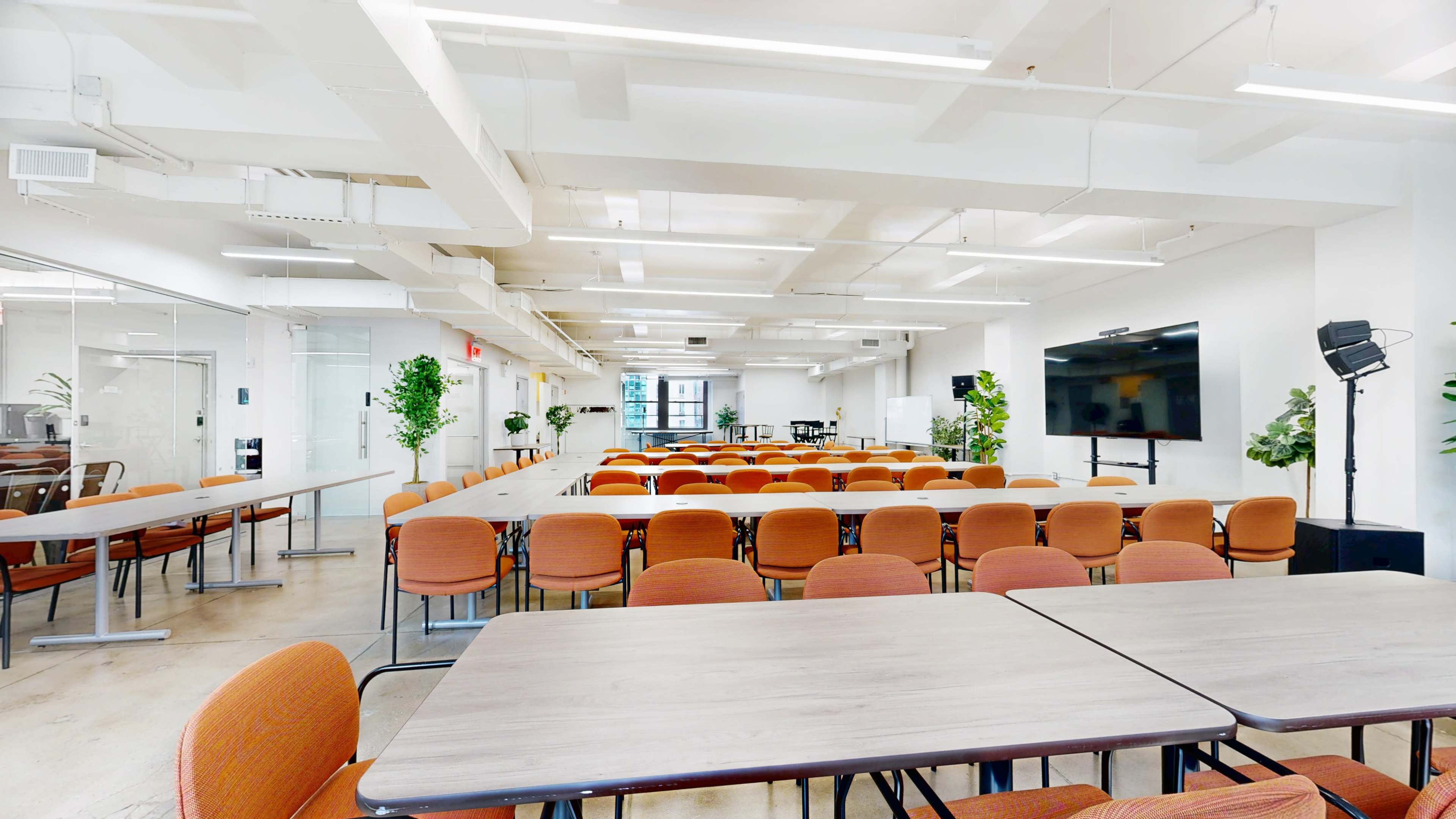 A conference room features multiple rows of orange chairs arranged around several tables in a well-lit, modern setting.