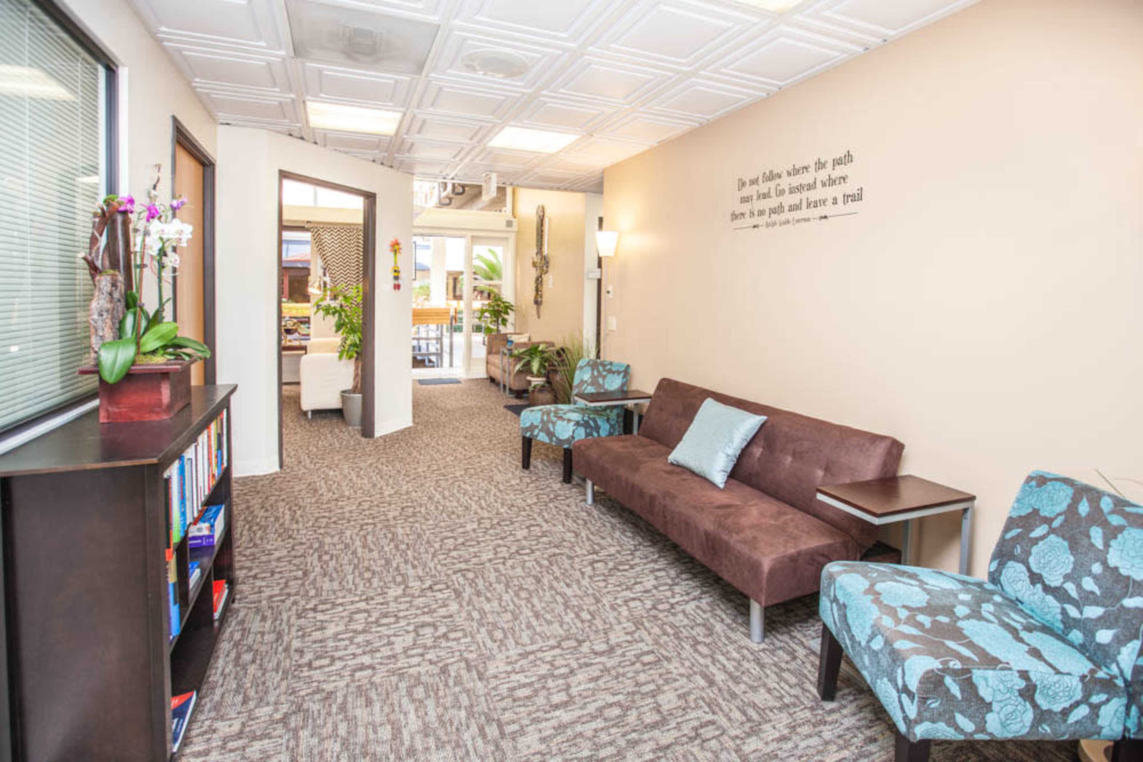 The image shows a well-lit hallway with seating and a bookshelf, leading to an open area decorated with plants and artwork.