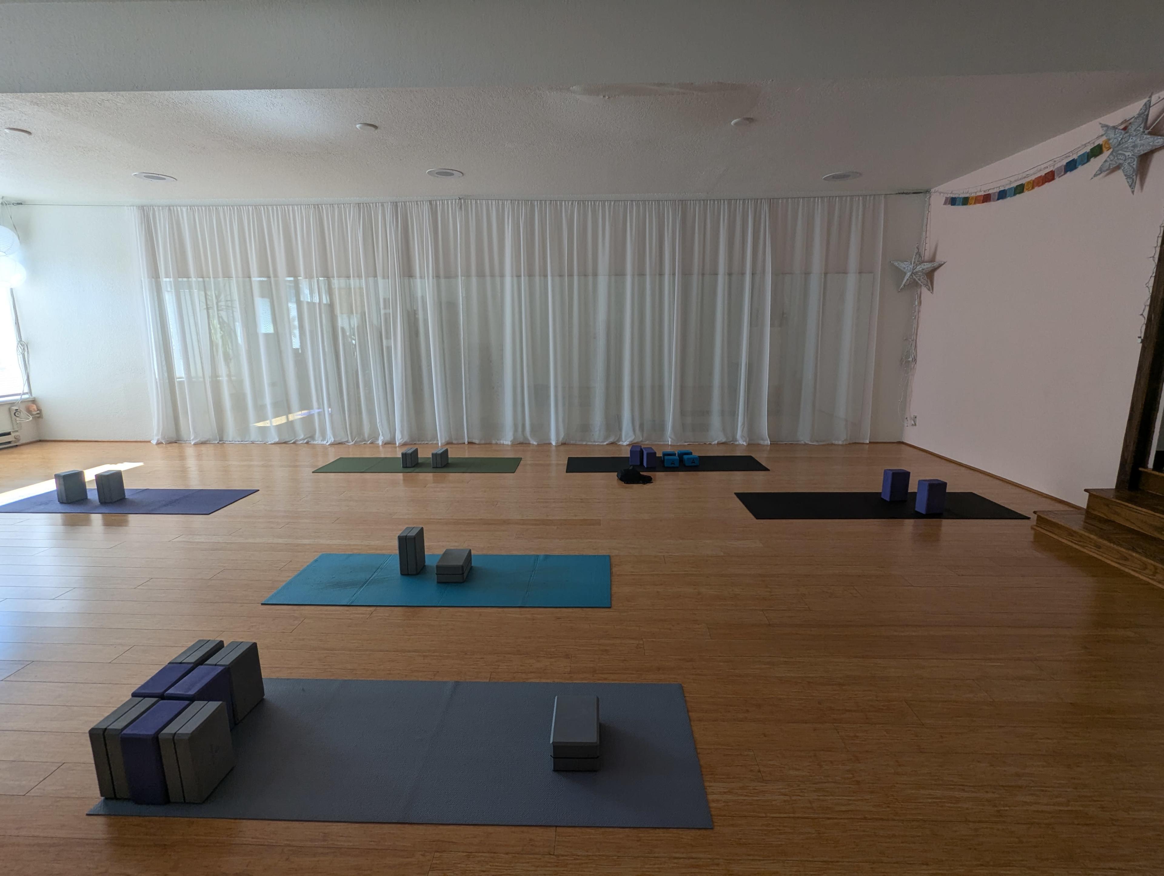 A spacious yoga studio with several mats arranged on a bamboo floor, accompanied by yoga blocks and props, and large sheer curtains in the background.