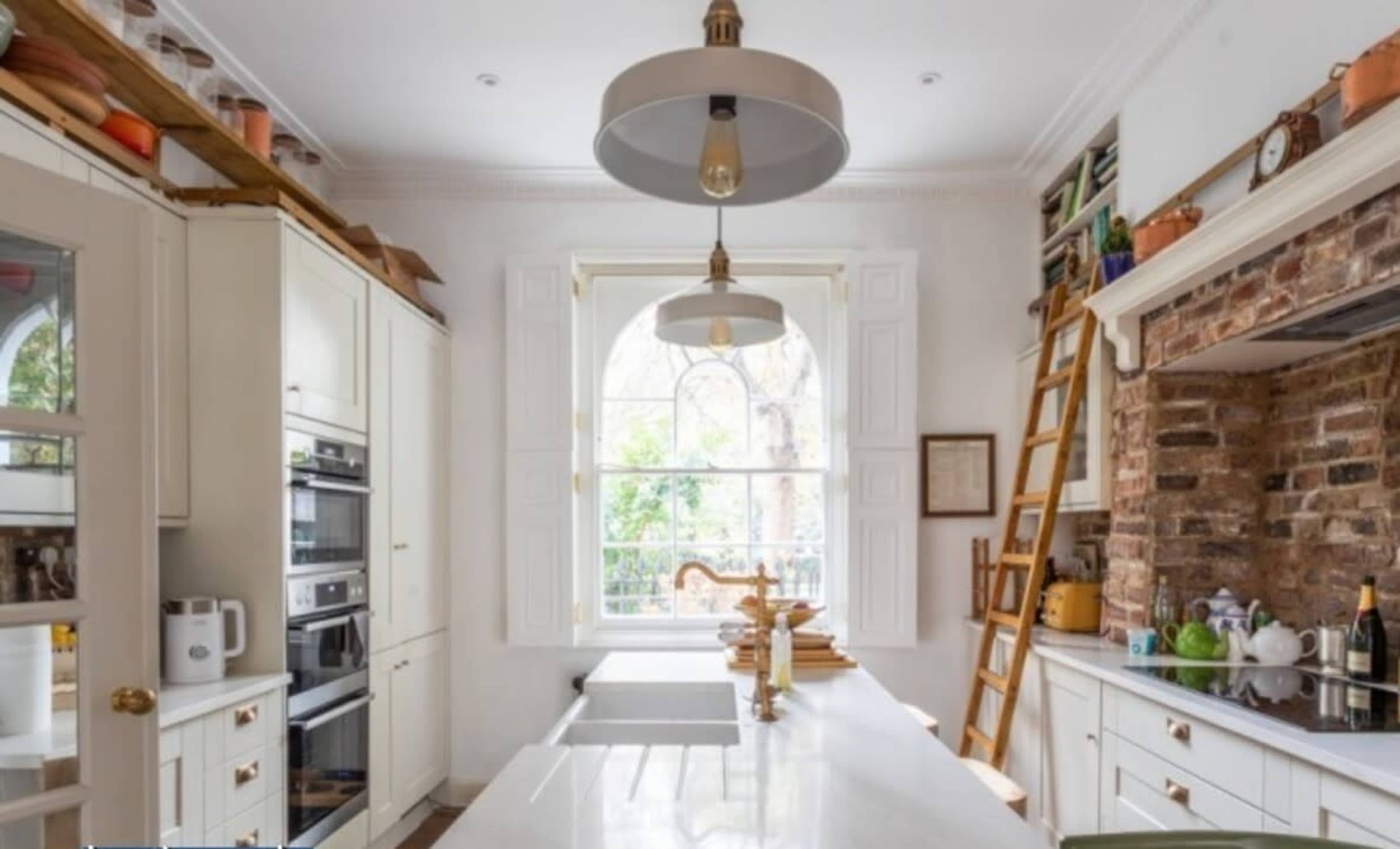 The kitchen features white cabinetry, a large marble island, and a brick accent wall, with natural light streaming through a large arched window.