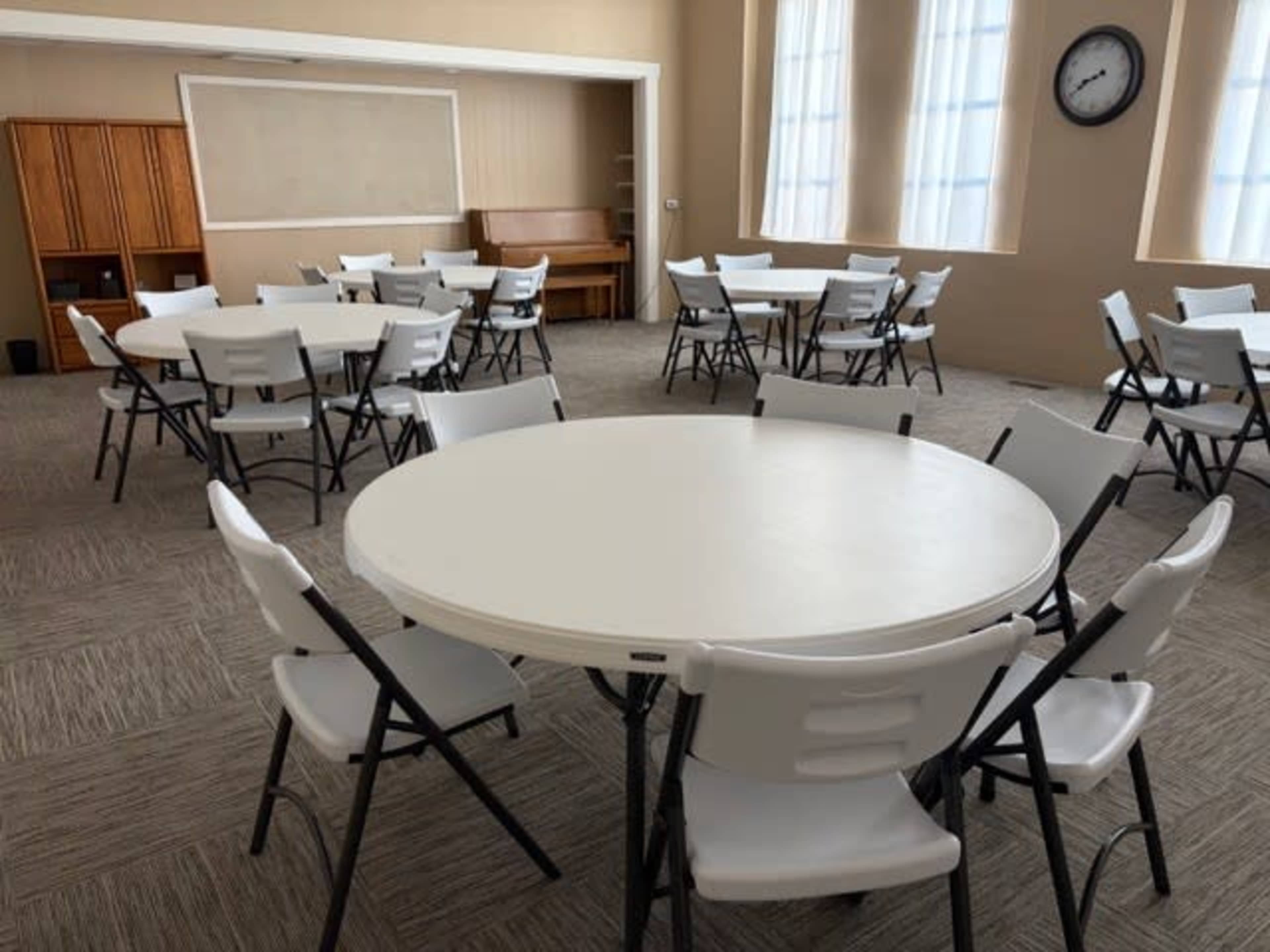 The image shows a room arranged with several round tables and chairs set up for an event, with a clock on the wall and wooden furniture in the background.