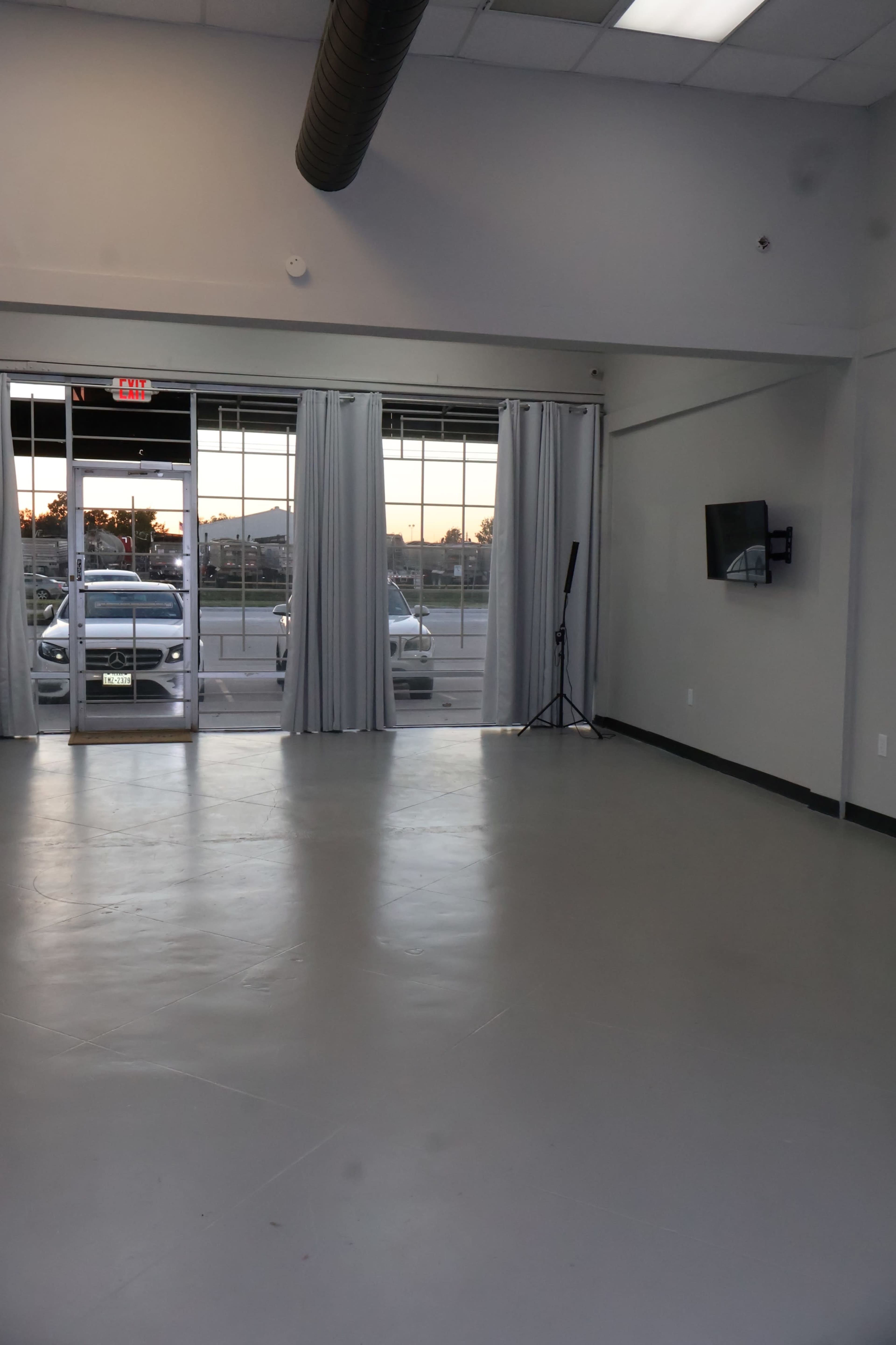 The image shows an empty room with a smooth, grey floor, large windows revealing two cars outside, and a television mounted on the wall.