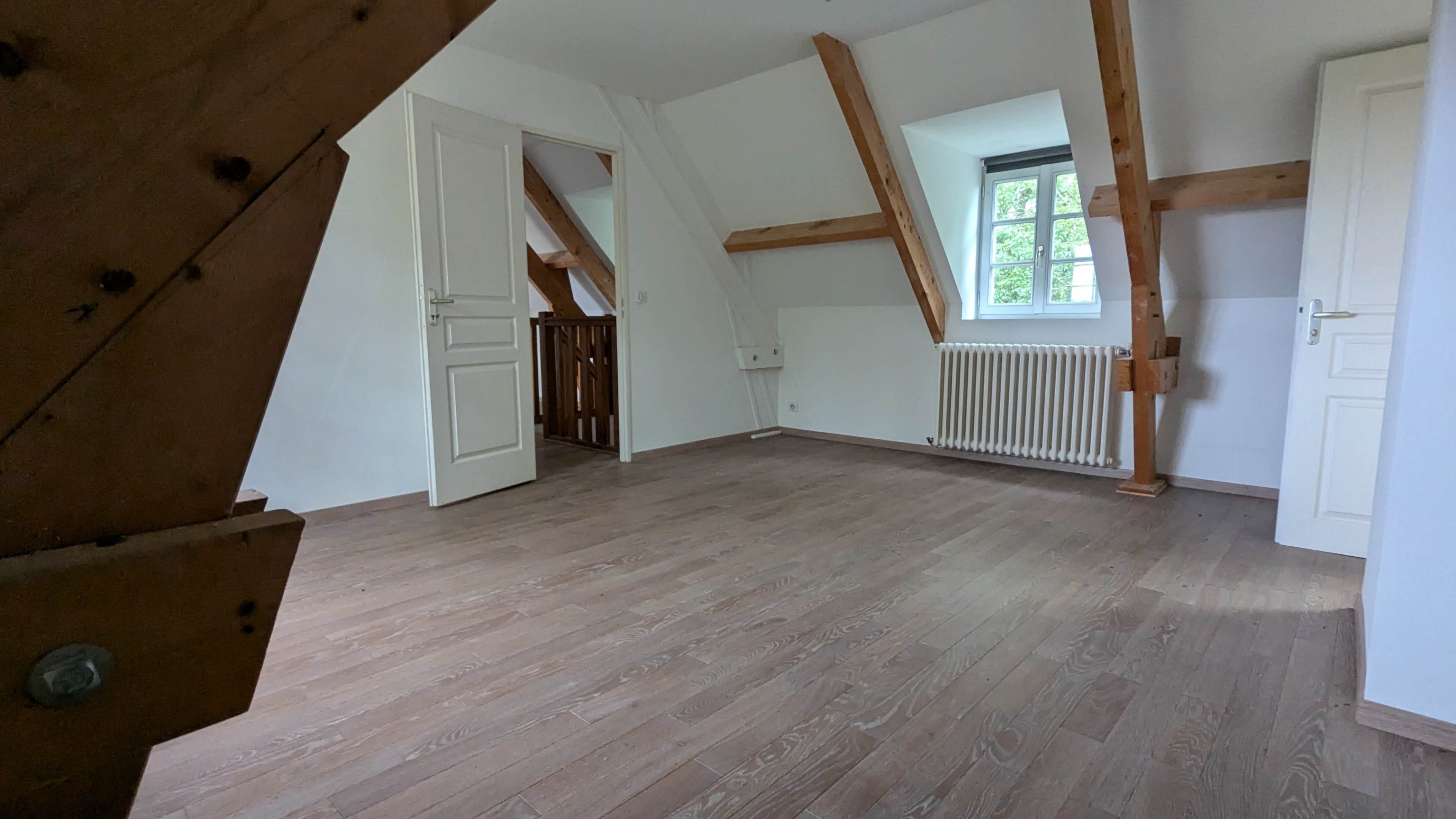 The image shows a spacious, empty room with wooden beams, light-colored walls, and a wooden floor, featuring a window and a door leading to another area.