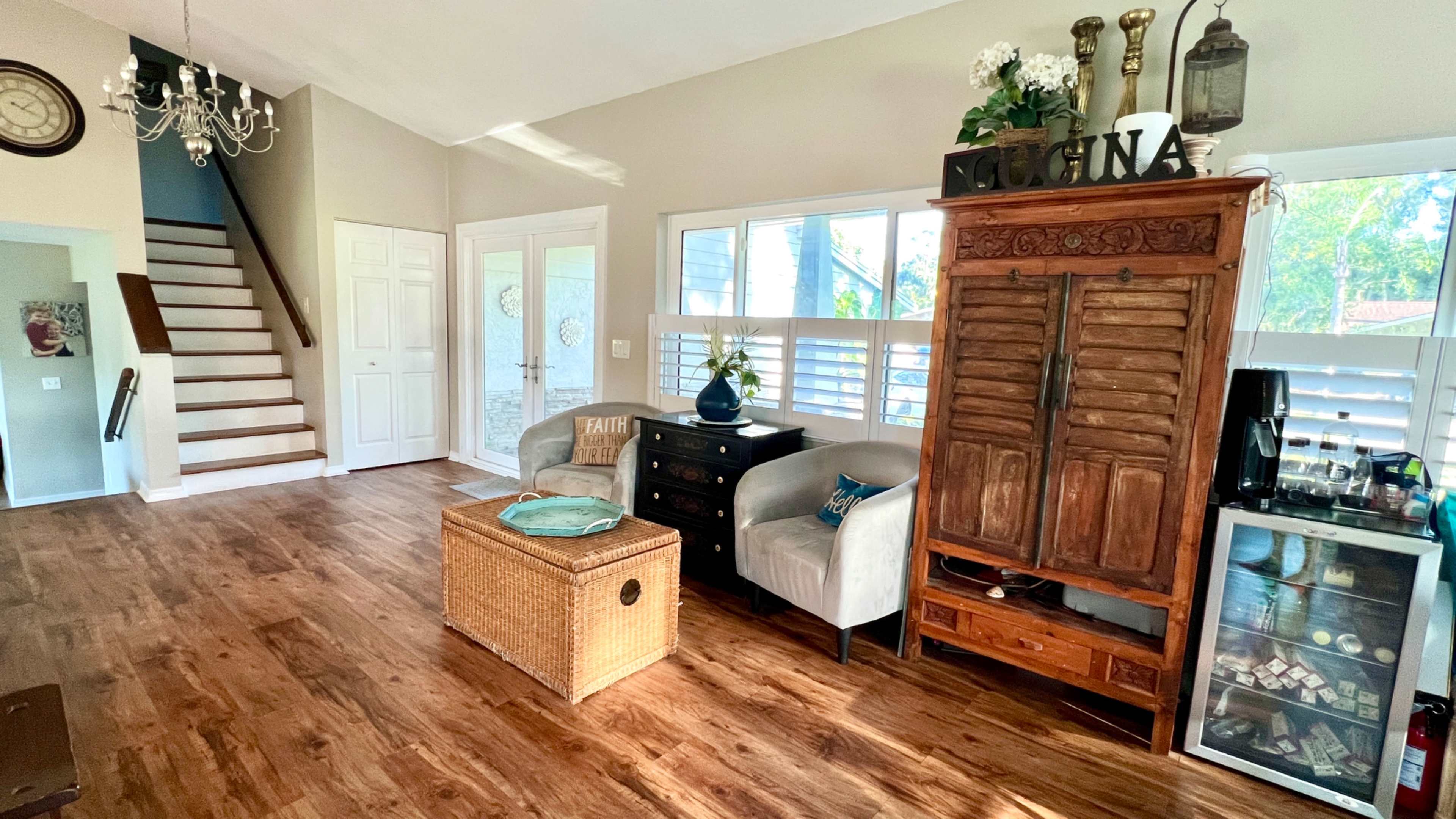 The image shows a living area with wooden flooring featuring a seating area, a decorative cabinet, a small wicker storage basket, and a staircase leading to an upper level.