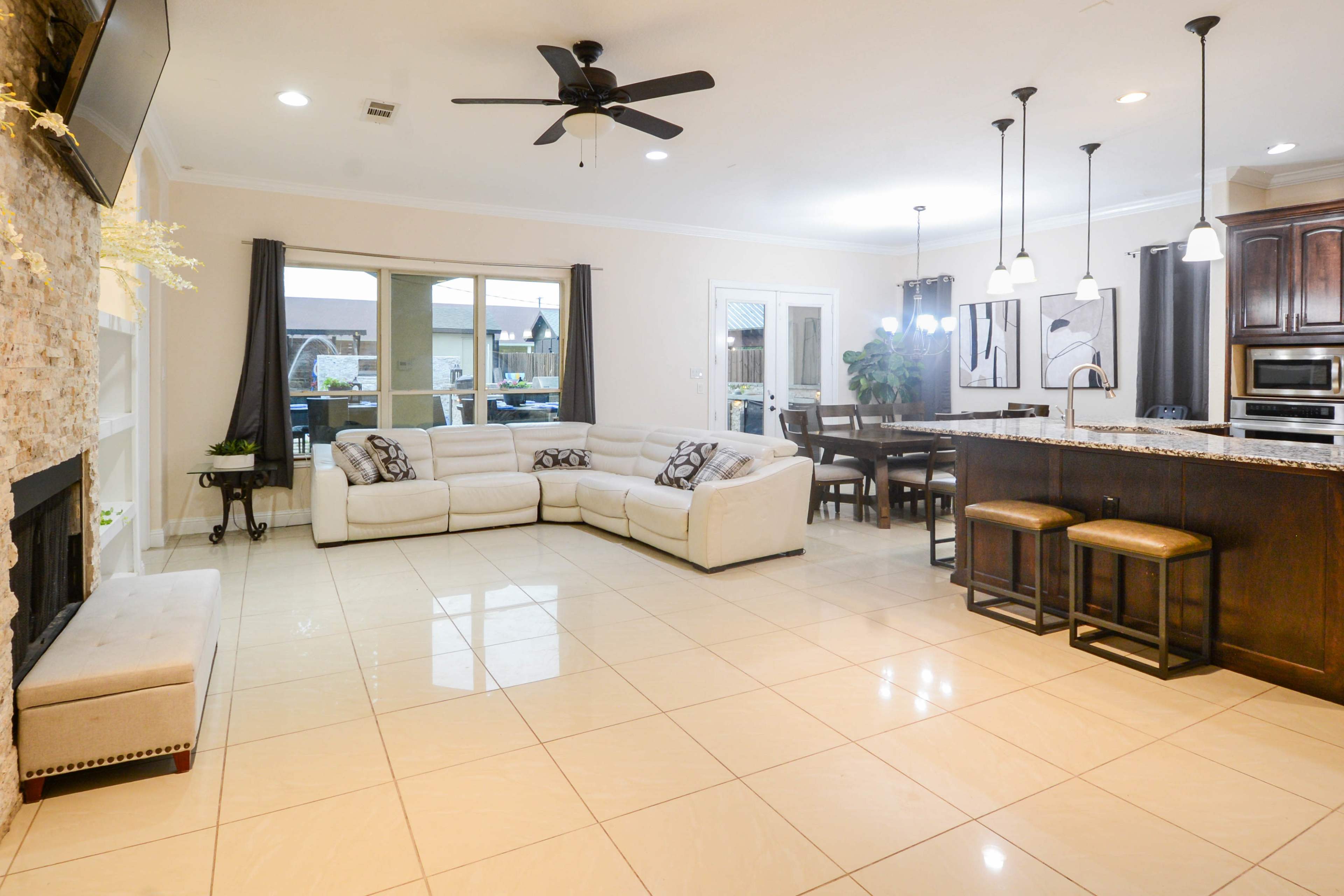 The image shows a spacious living area and kitchen with modern furnishings, featuring a sectional sofa, dining table, and a large window.