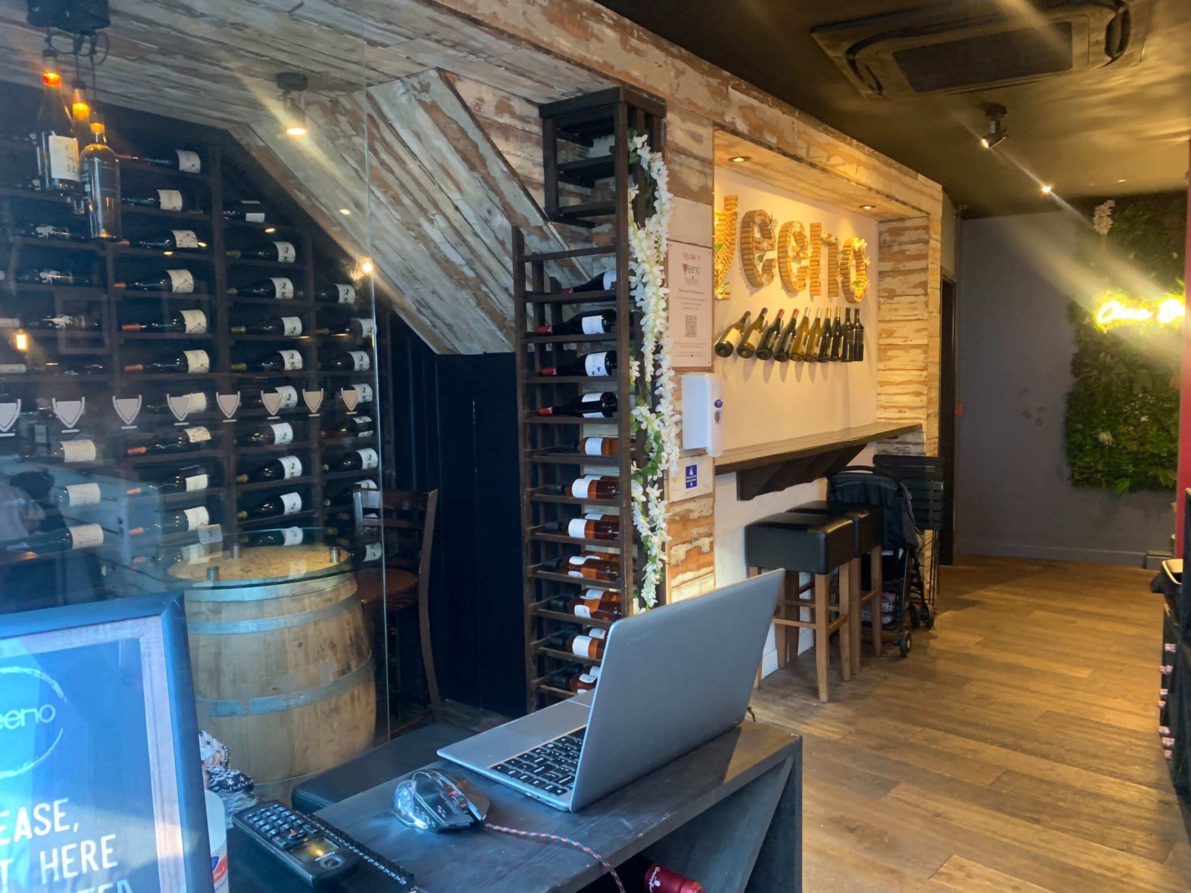 A wine bar interior with a wooden wall, wine bottles neatly arranged on shelves, and a laptop on a black counter.