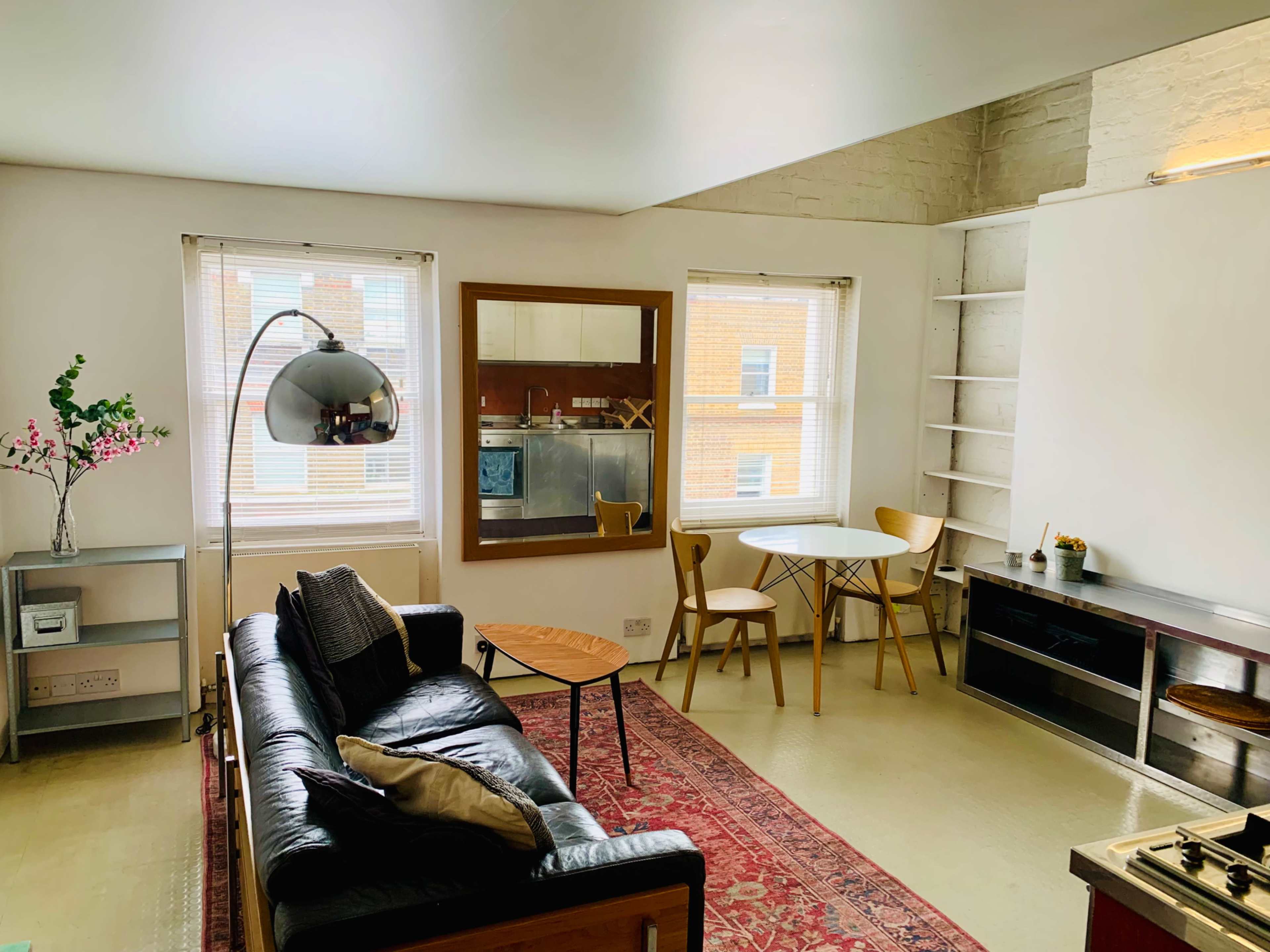 The image shows a living room featuring a black leather sofa, a wooden coffee table, a small dining area with two chairs, and a kitchenette in the background.