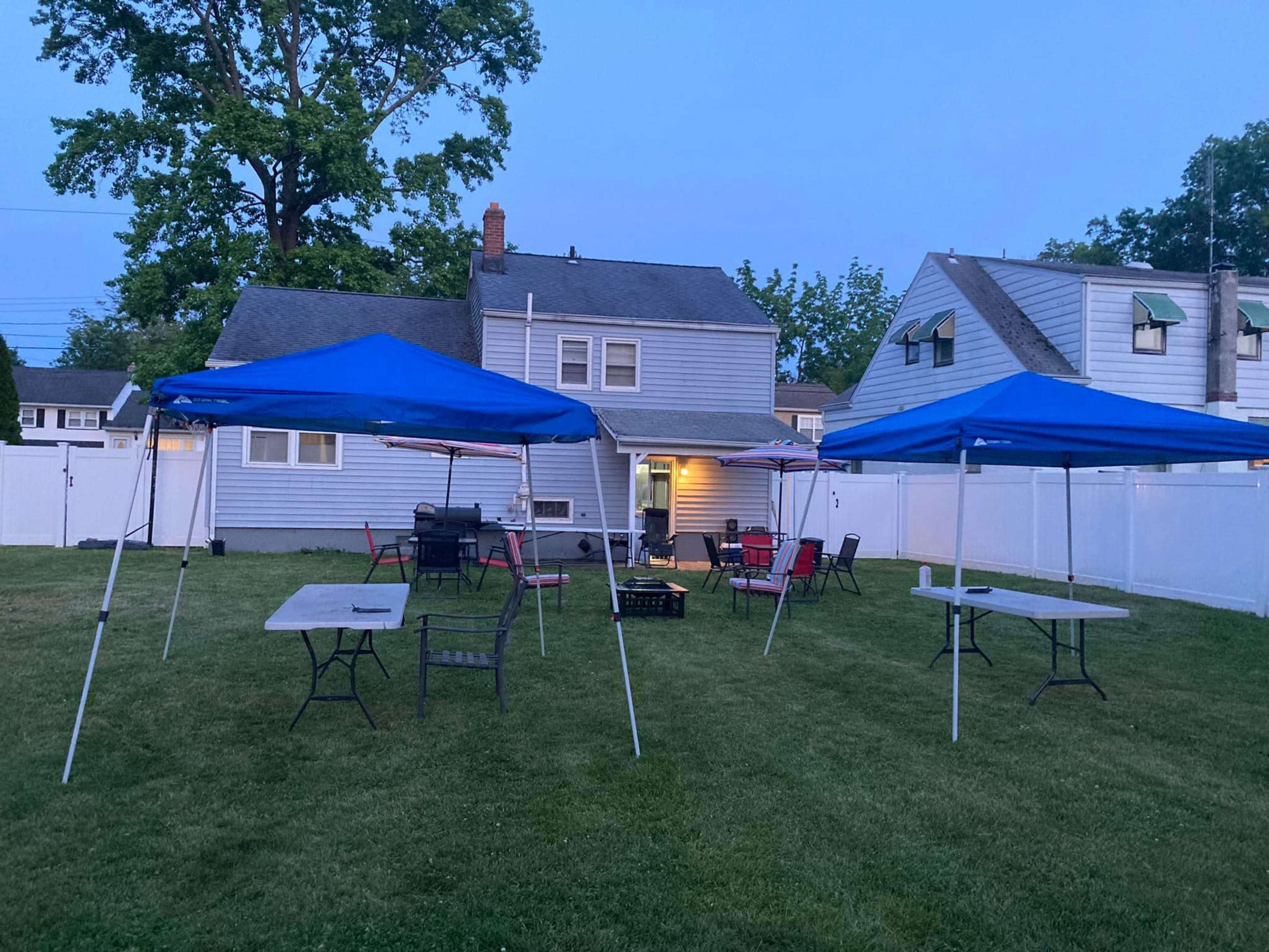 A backyard with two blue party tents set up on a grassy area in front of a house.