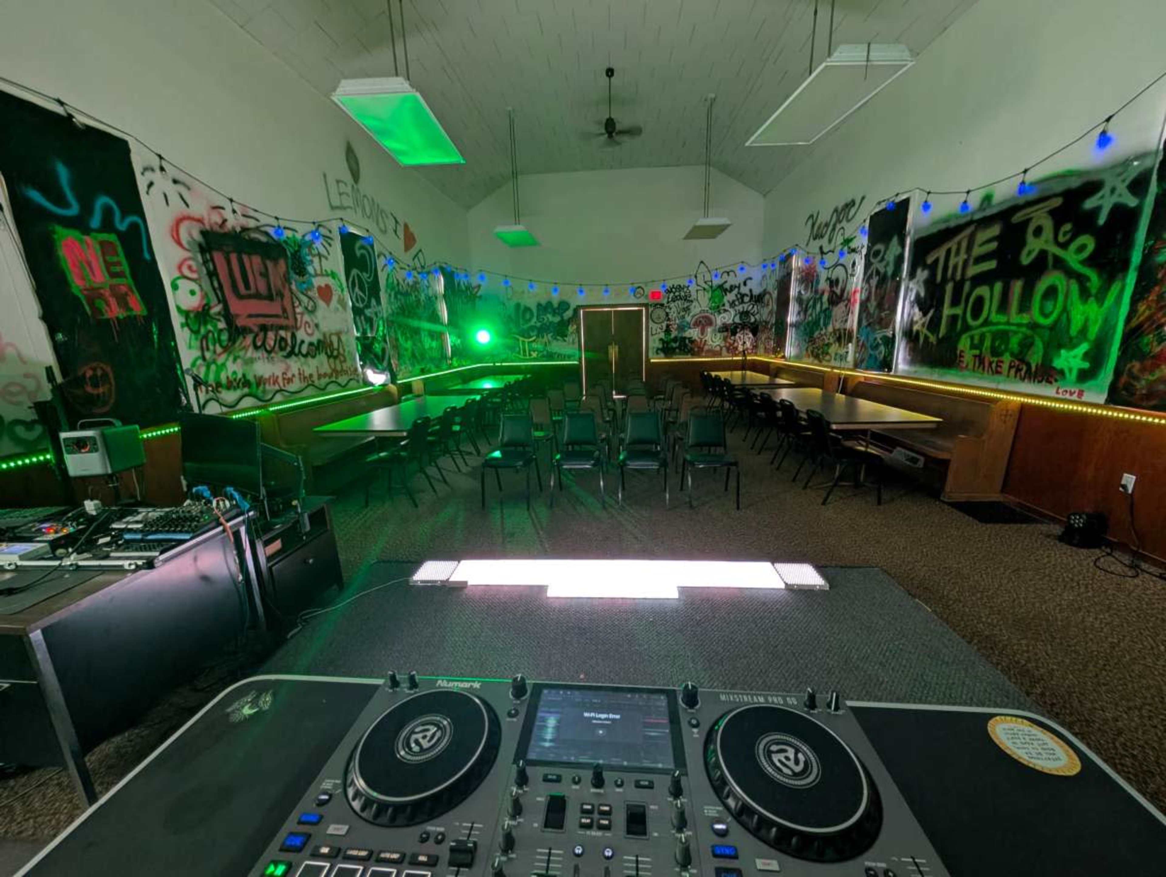 The image shows a dimly lit room with graffiti-covered walls, chairs arranged in rows, and a DJ setup positioned in the foreground.