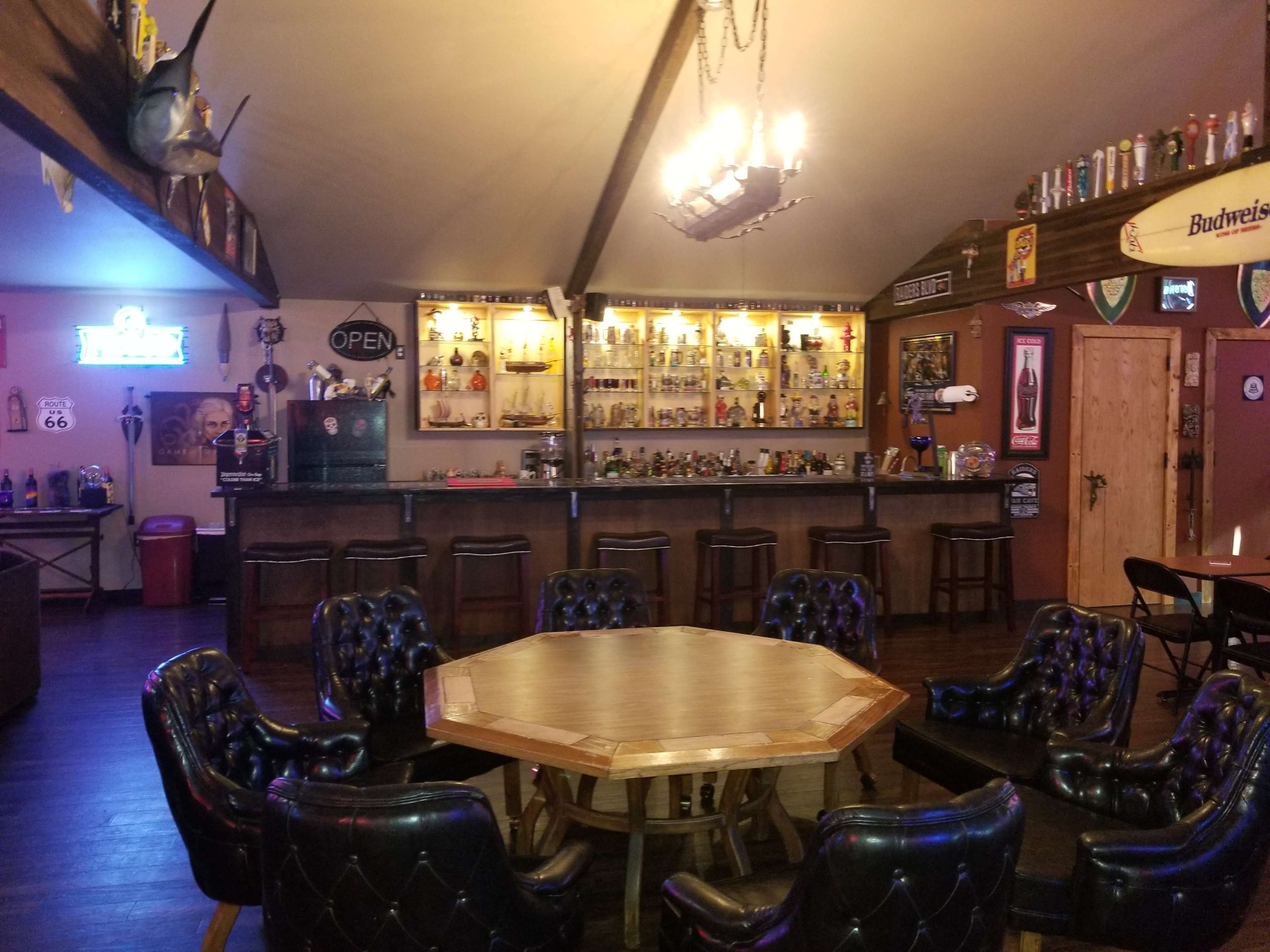A rustic bar area with a wooden counter, several bar stools, a display of bottles on shelves, and a large octagonal table surrounded by black upholstered chairs.