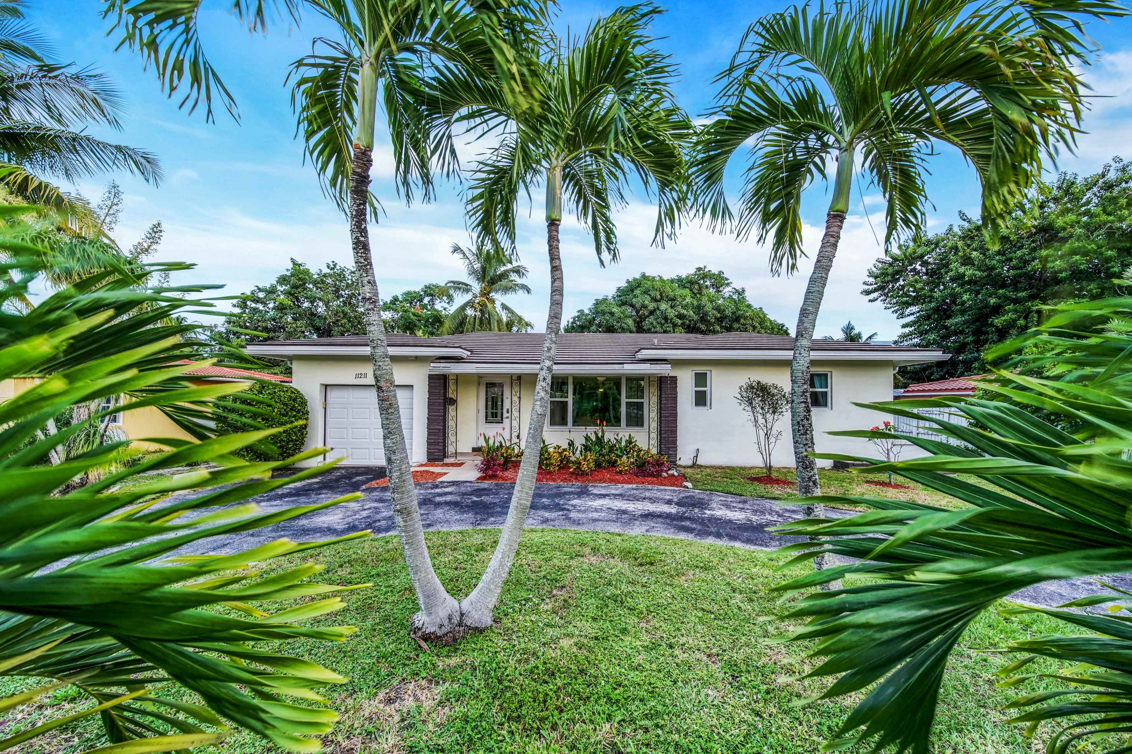 A single-story house with palm trees in the foreground and a driveway leading to the entrance is visible.