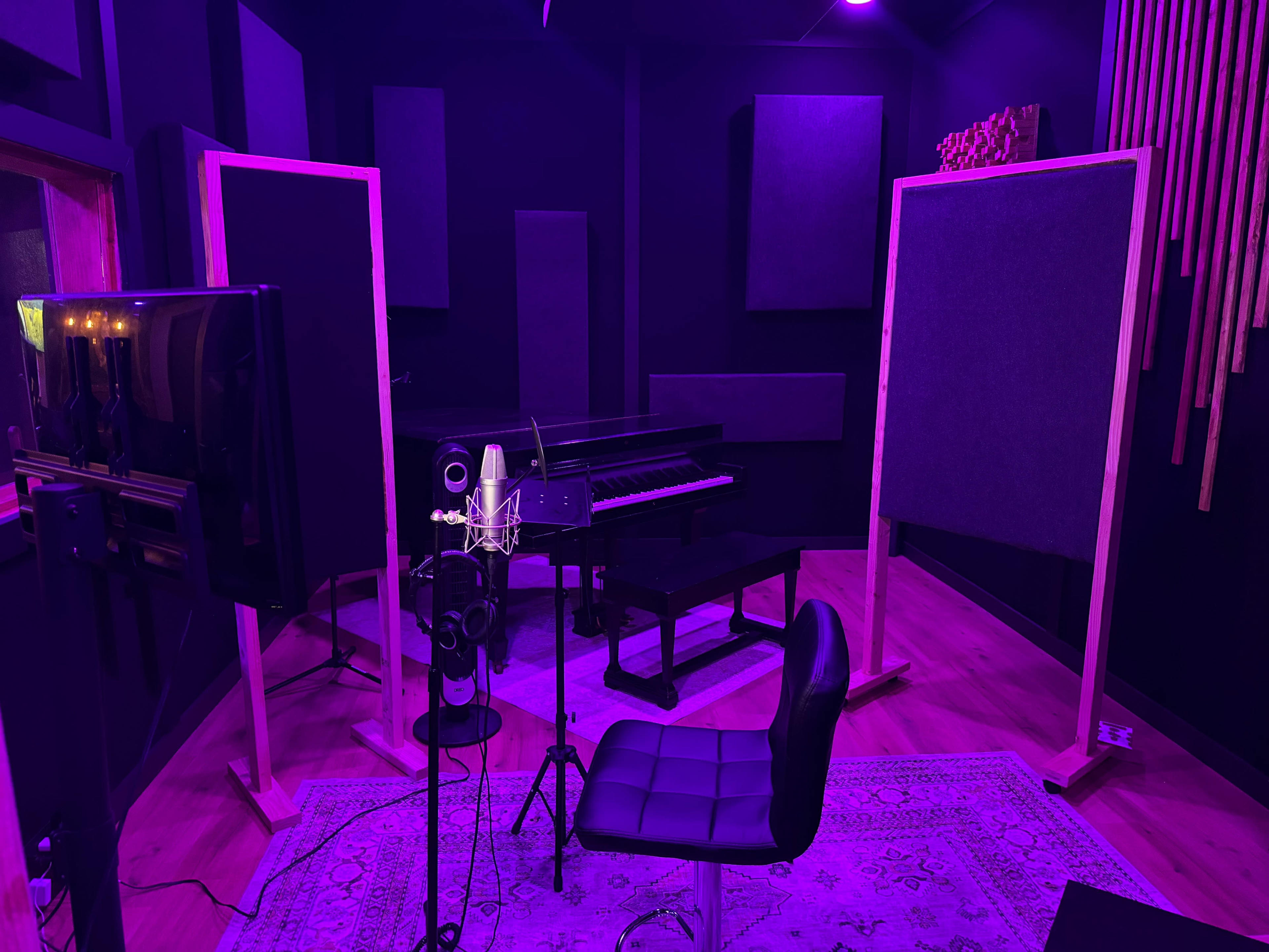 The image shows a dimly lit recording studio with purple lighting, featuring a microphone setup, a black piano, and soundproof panels.
