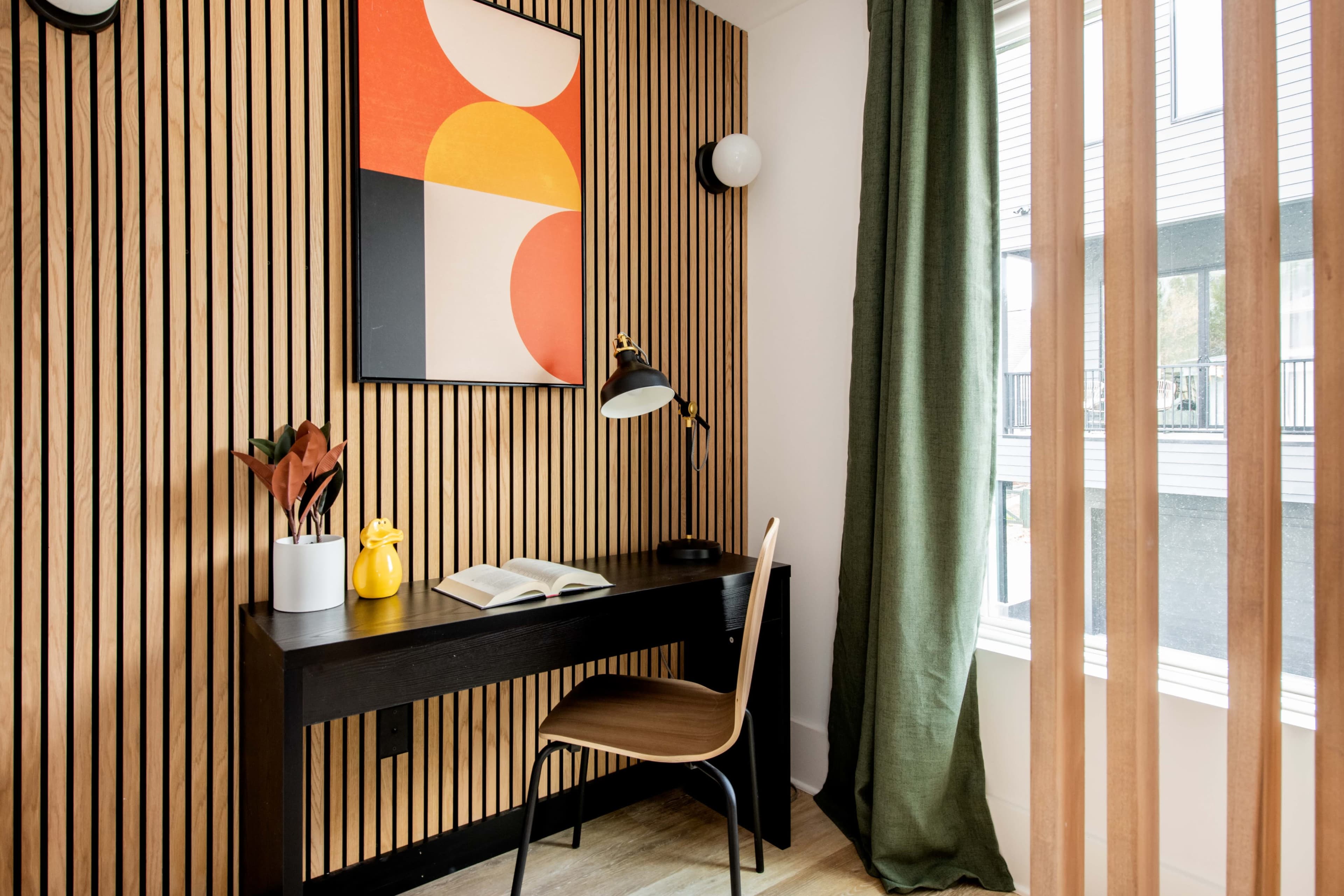 The image shows a small workspace with a black desk, a chair, an open book, a lamp, and a colorful abstract artwork on the wall, surrounded by wooden wall panels and green curtains.