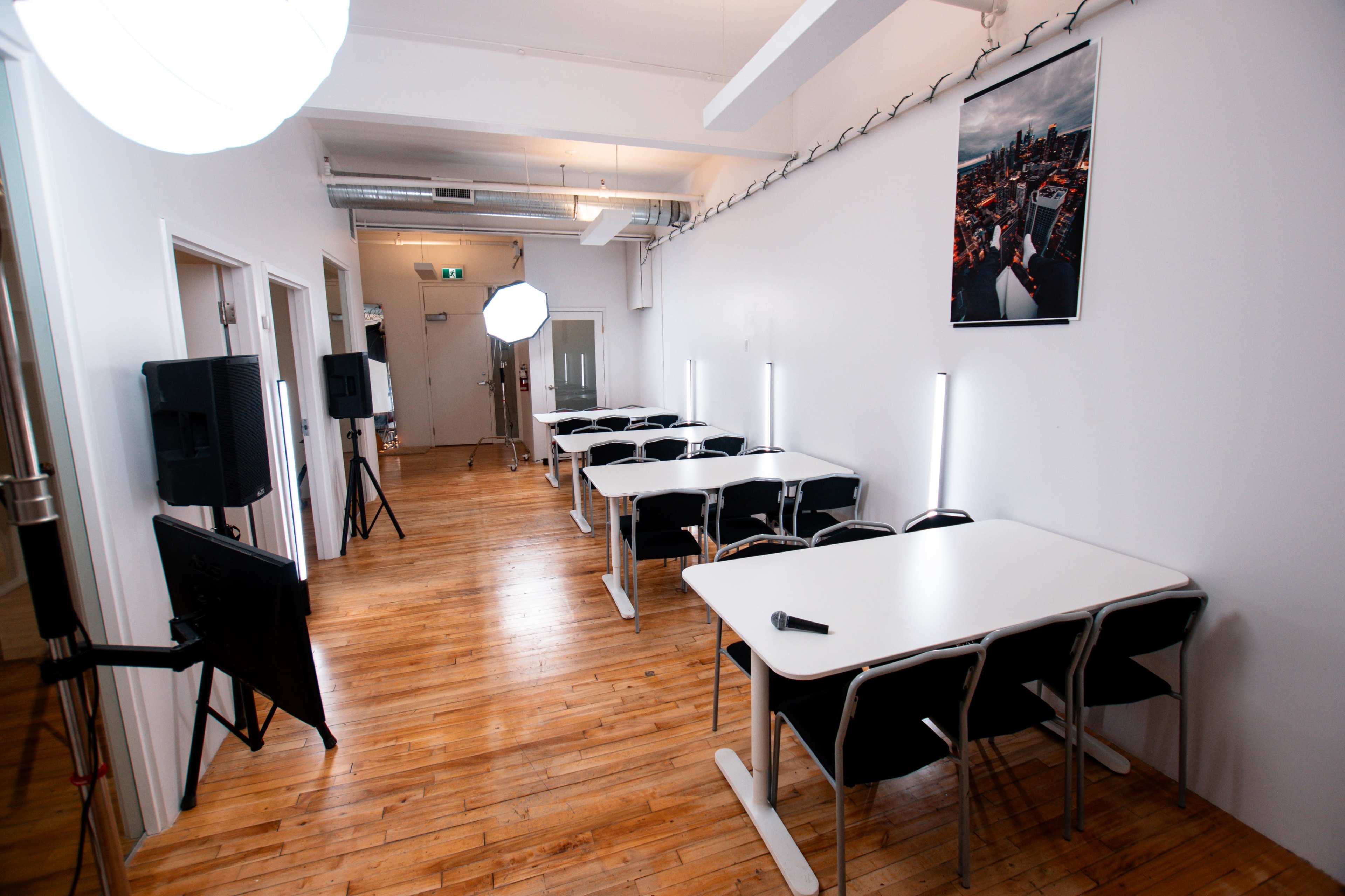 The image shows a modern, well-lit room with wooden floors, featuring several white tables and black chairs arranged in rows, along with softbox lights and a large photo on the wall.