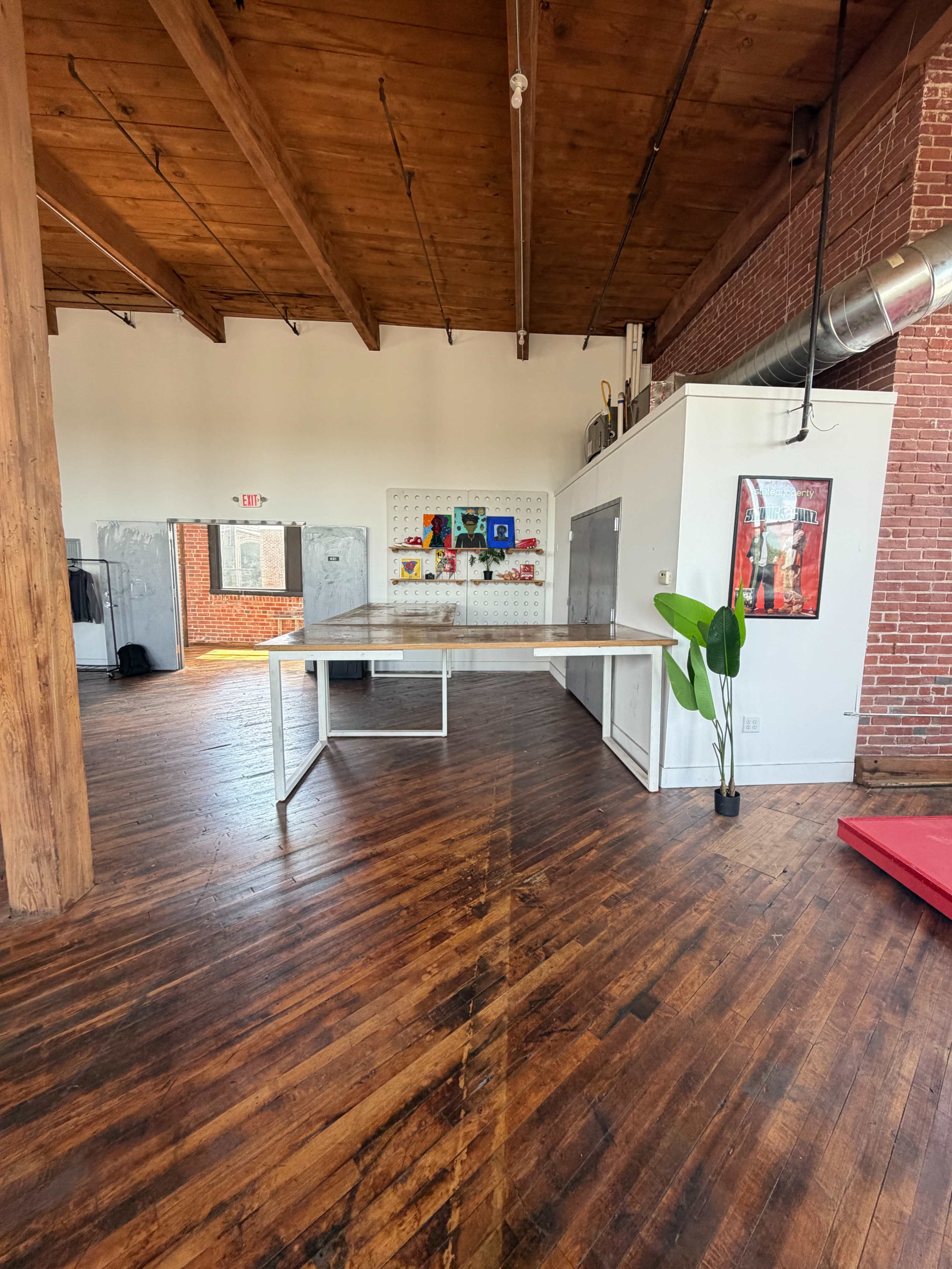 The image shows a spacious room with wooden floors, featuring a large white table in the center, and exposed brick walls in the background.