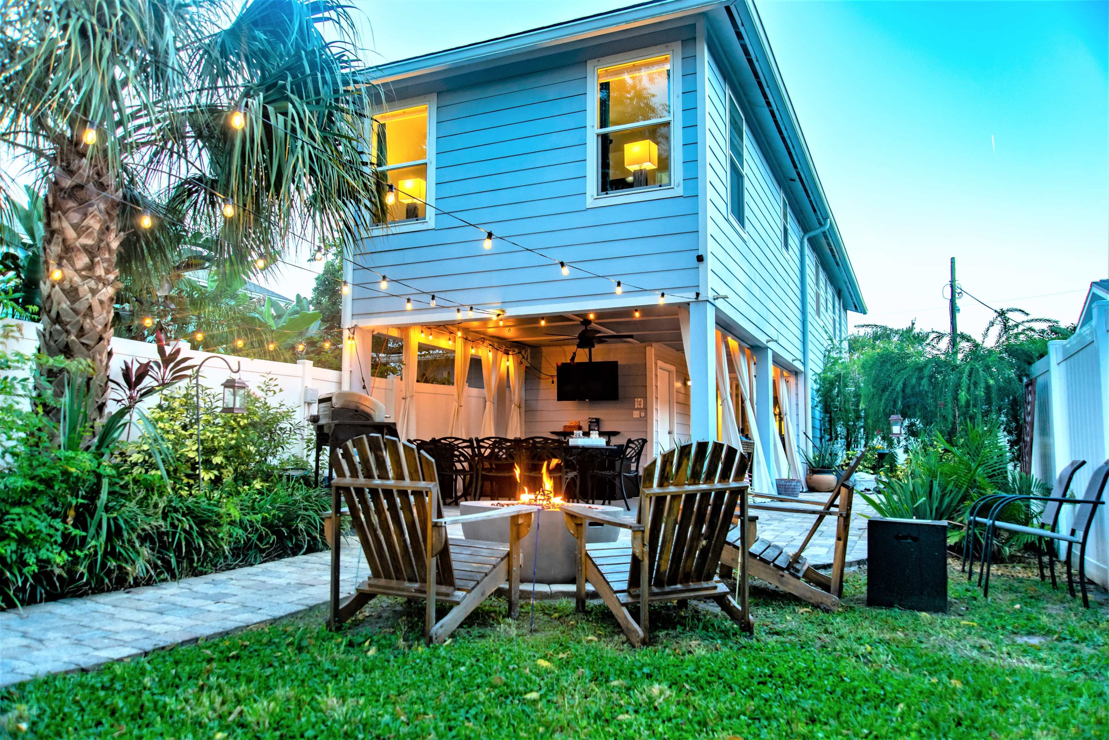 A backyard scene features a blue two-story house with string lights, an outdoor dining area, and a fire pit surrounded by wooden chairs.