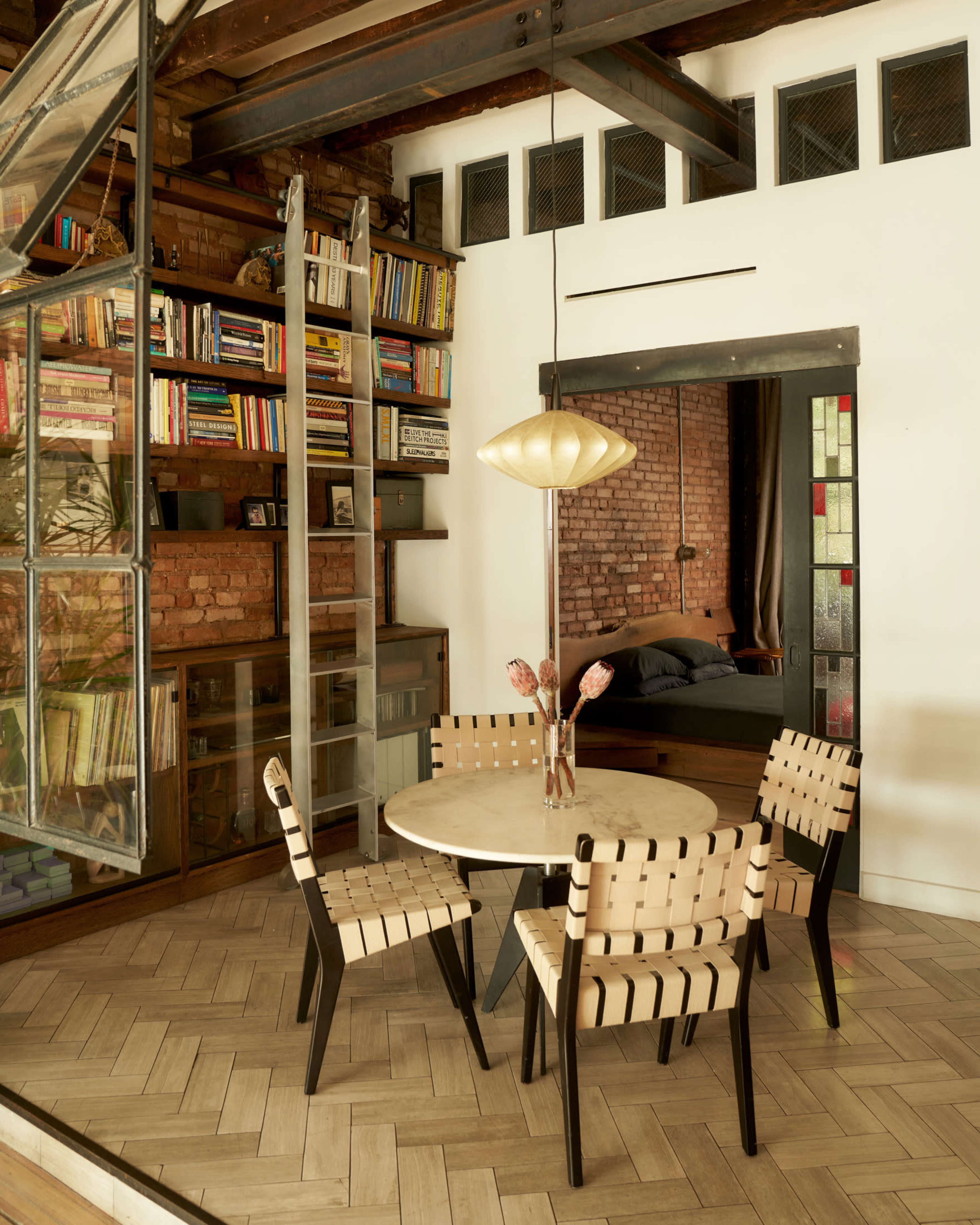 A round table with four wooden chairs is set in a contemporary room featuring a bookshelf filled with colorful books and exposed brick walls.