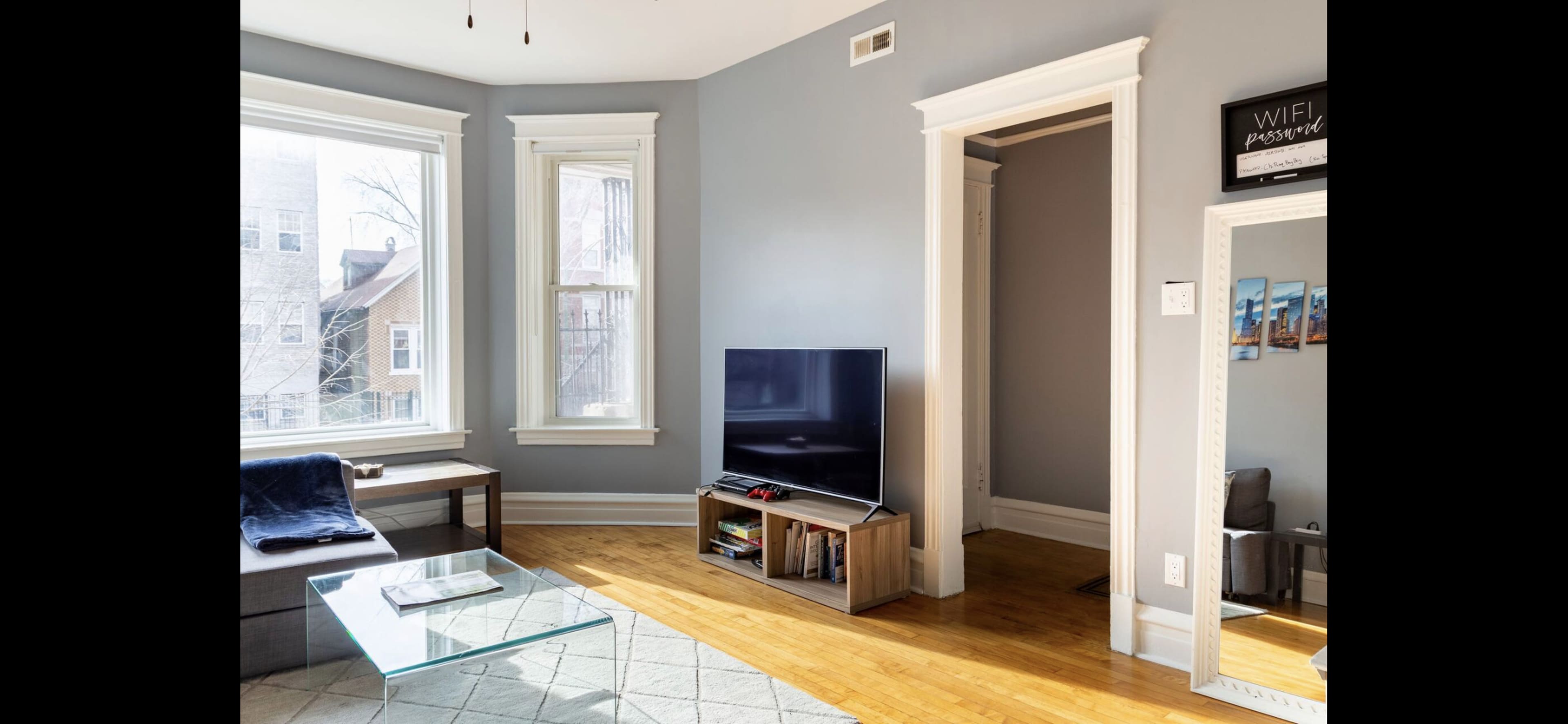 A living room features a modern television stand, a glass coffee table, and large windows allowing natural light to enter.