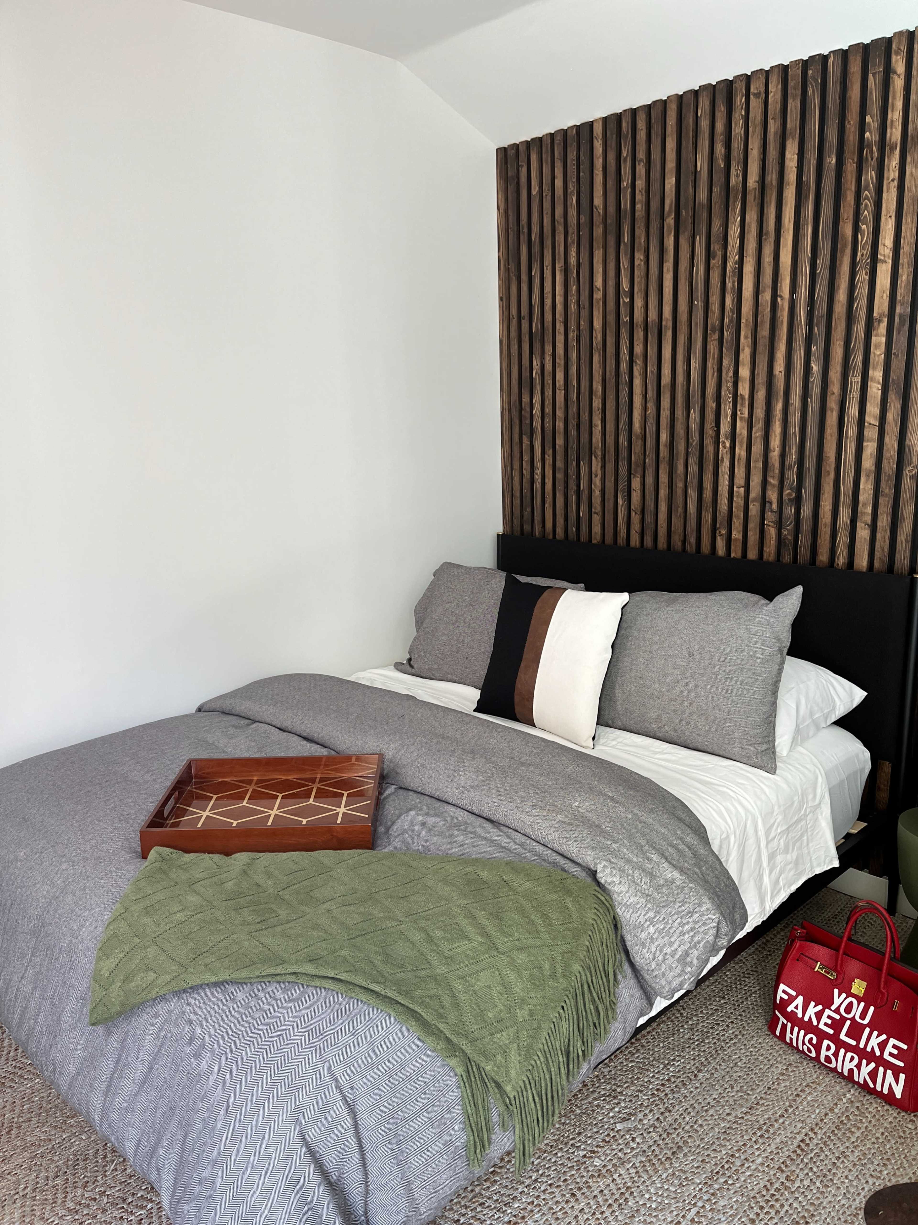 A neatly made bed with gray bedding and decorative pillows is positioned against a wooden accent wall, accompanied by a green throw and a wooden tray on the bed.