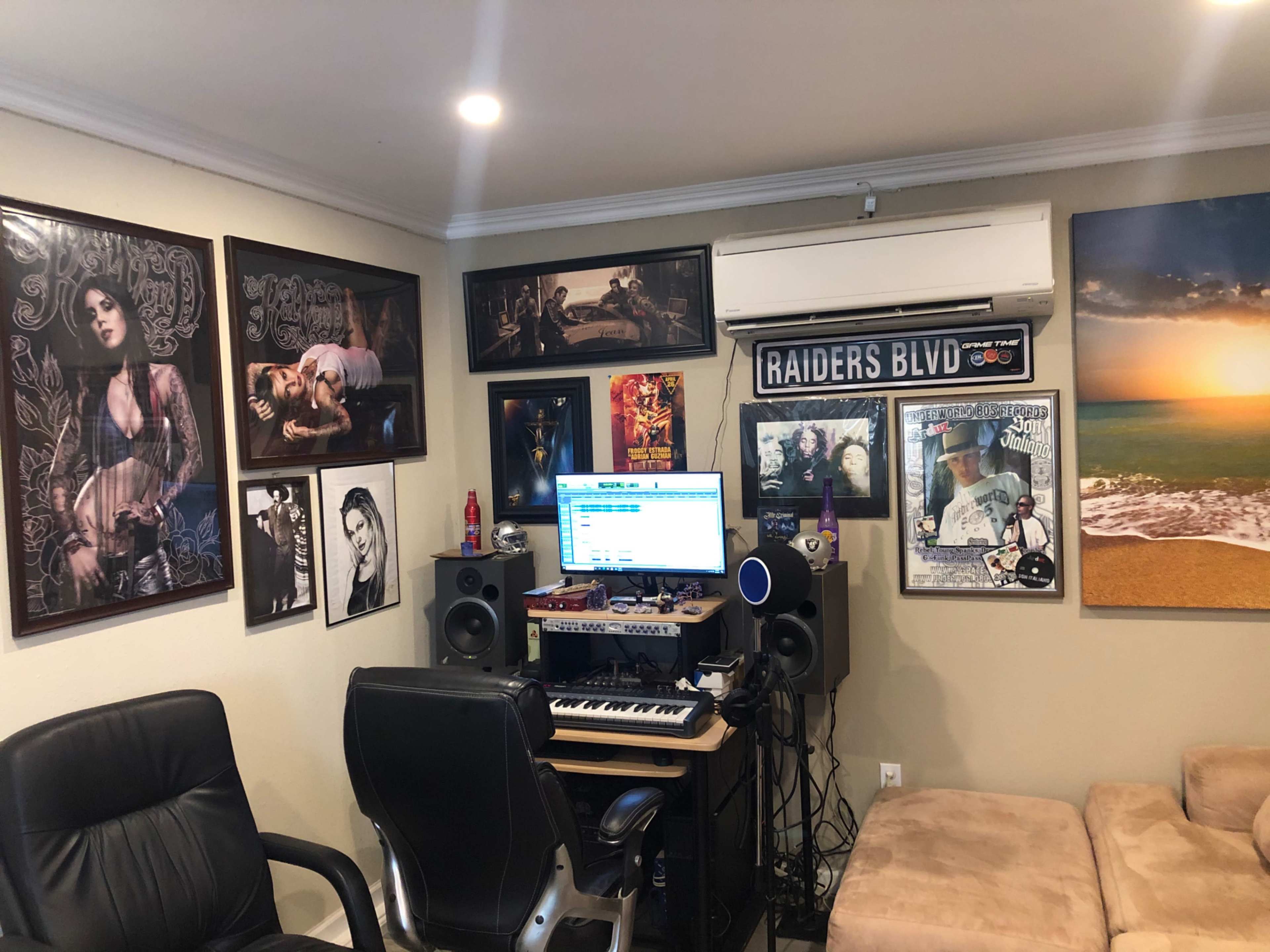A home recording studio with a computer, keyboards, speakers, and numerous framed posters on the walls.