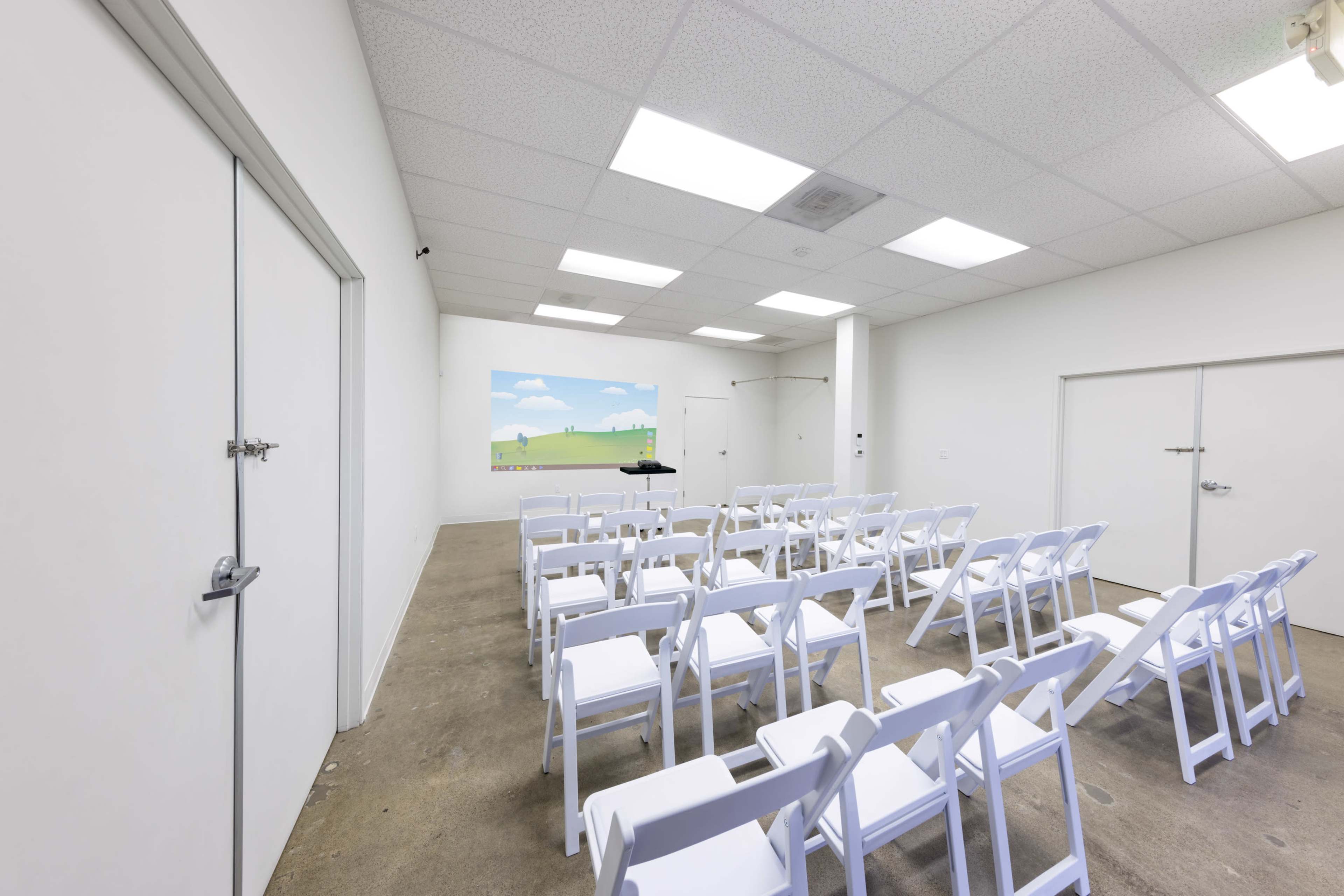 A spacious, empty room with white folding chairs arranged in rows facing a screen displaying a simple landscape.