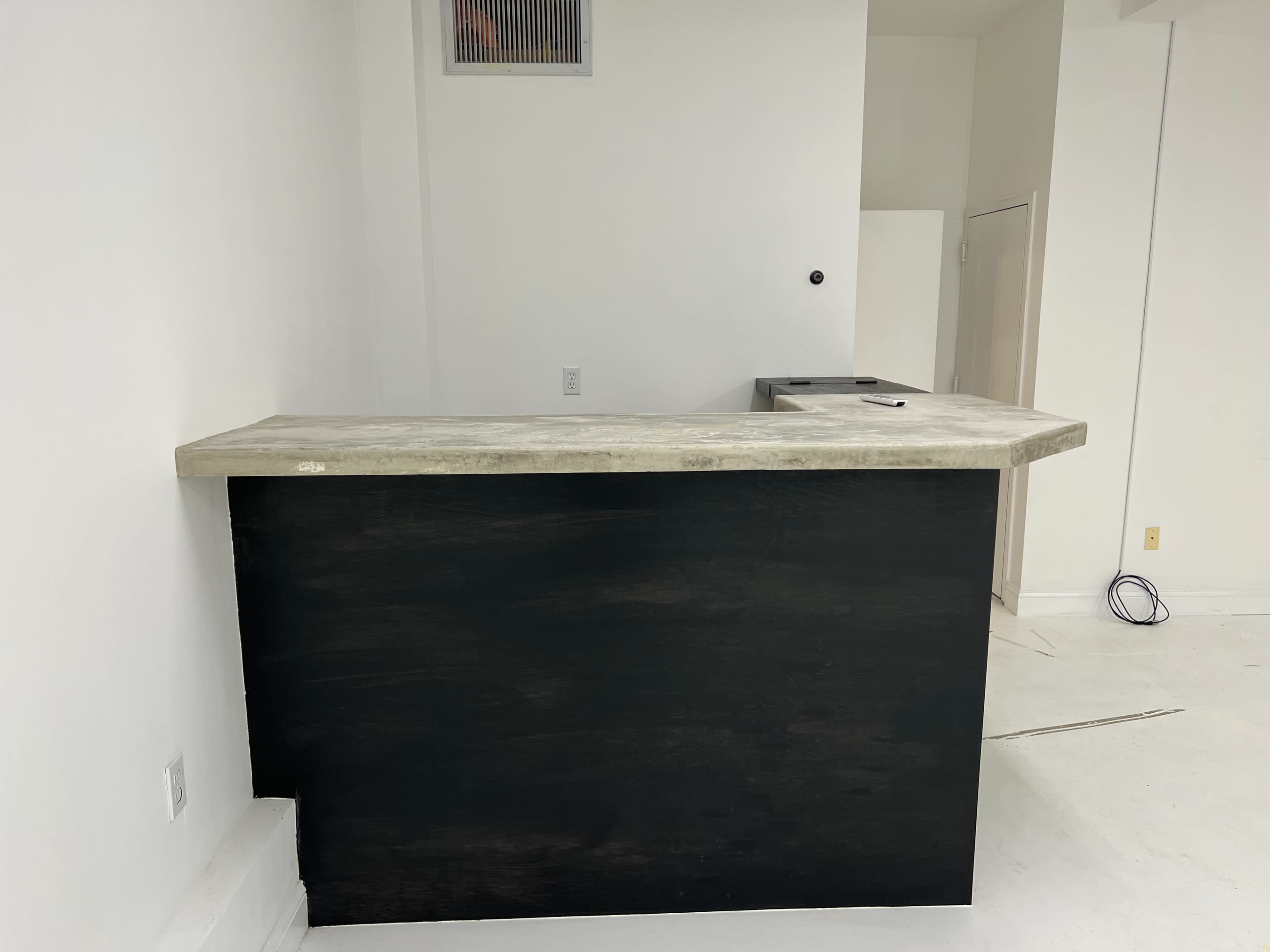 A minimalist reception counter with a concrete top and a dark wooden base in a white-walled space.