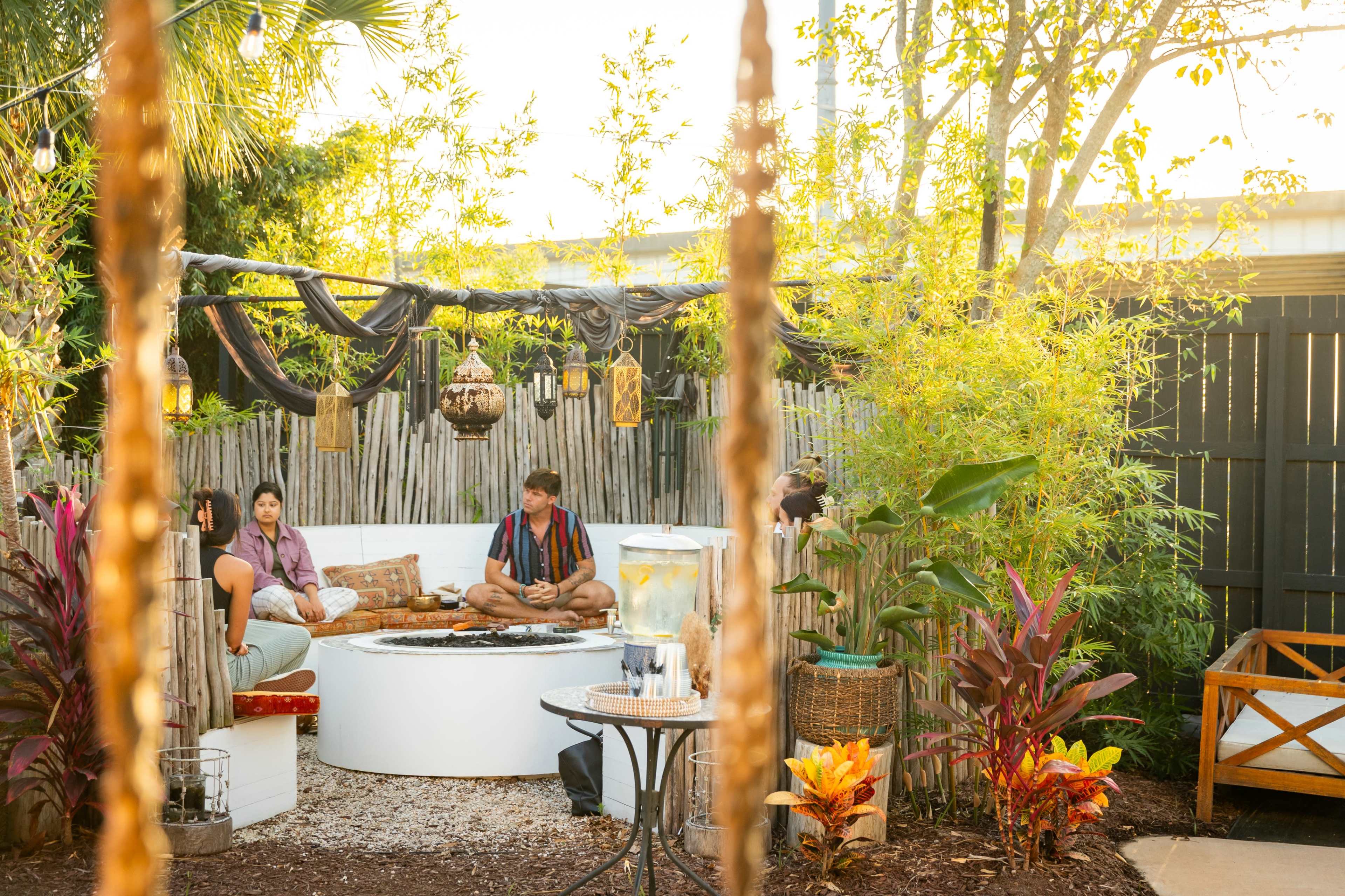 A group of three people sits around a circular fire pit in a backyard surrounded by plants and decorative elements.