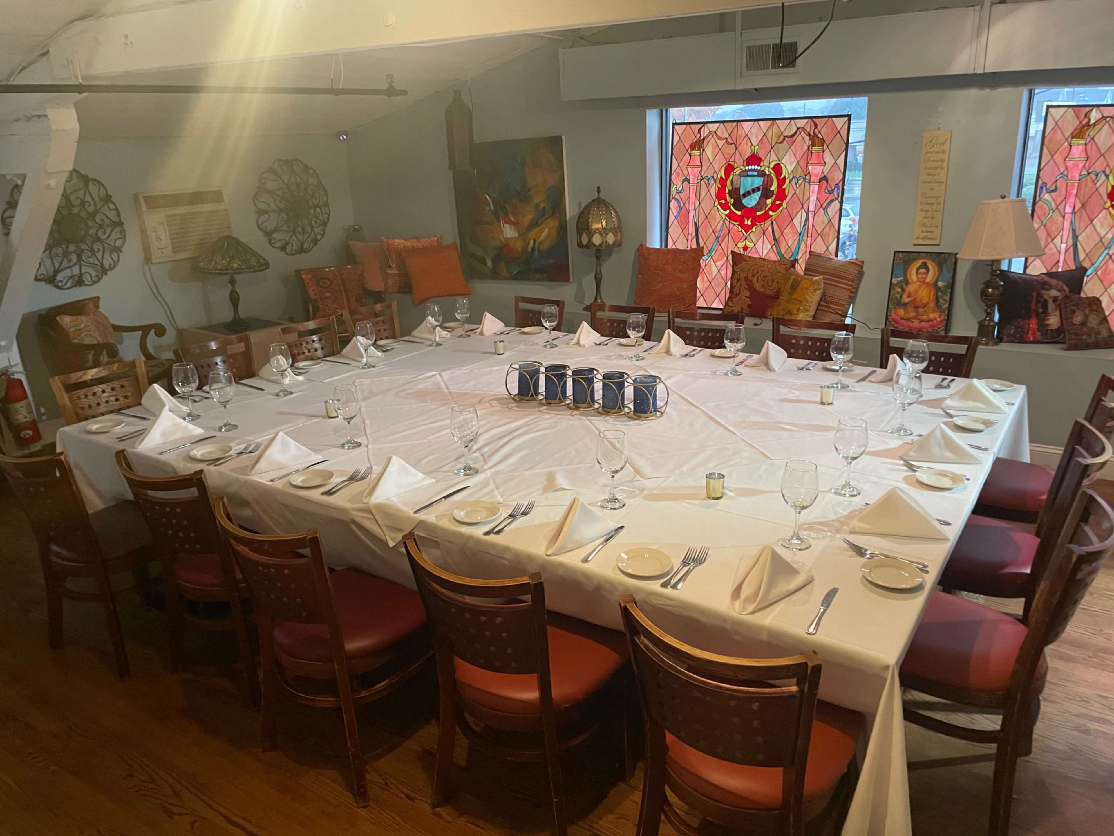 A large, elegantly set dining table with white linens and glassware is arranged in a room decorated with various artworks and stained glass windows.