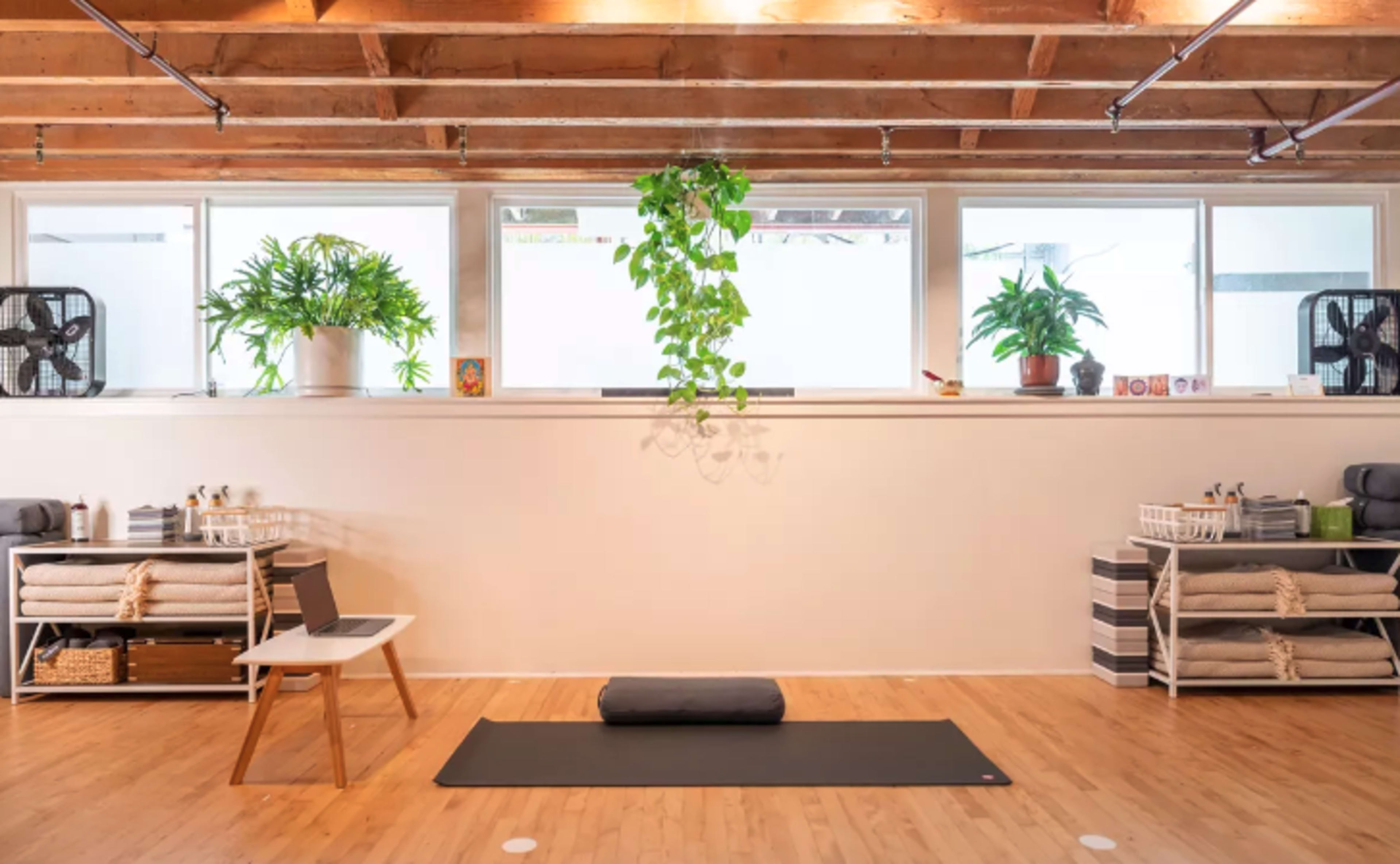 A yoga studio features a mat on the floor, potted plants on windowsills, and neatly organized shelves with blankets and props.