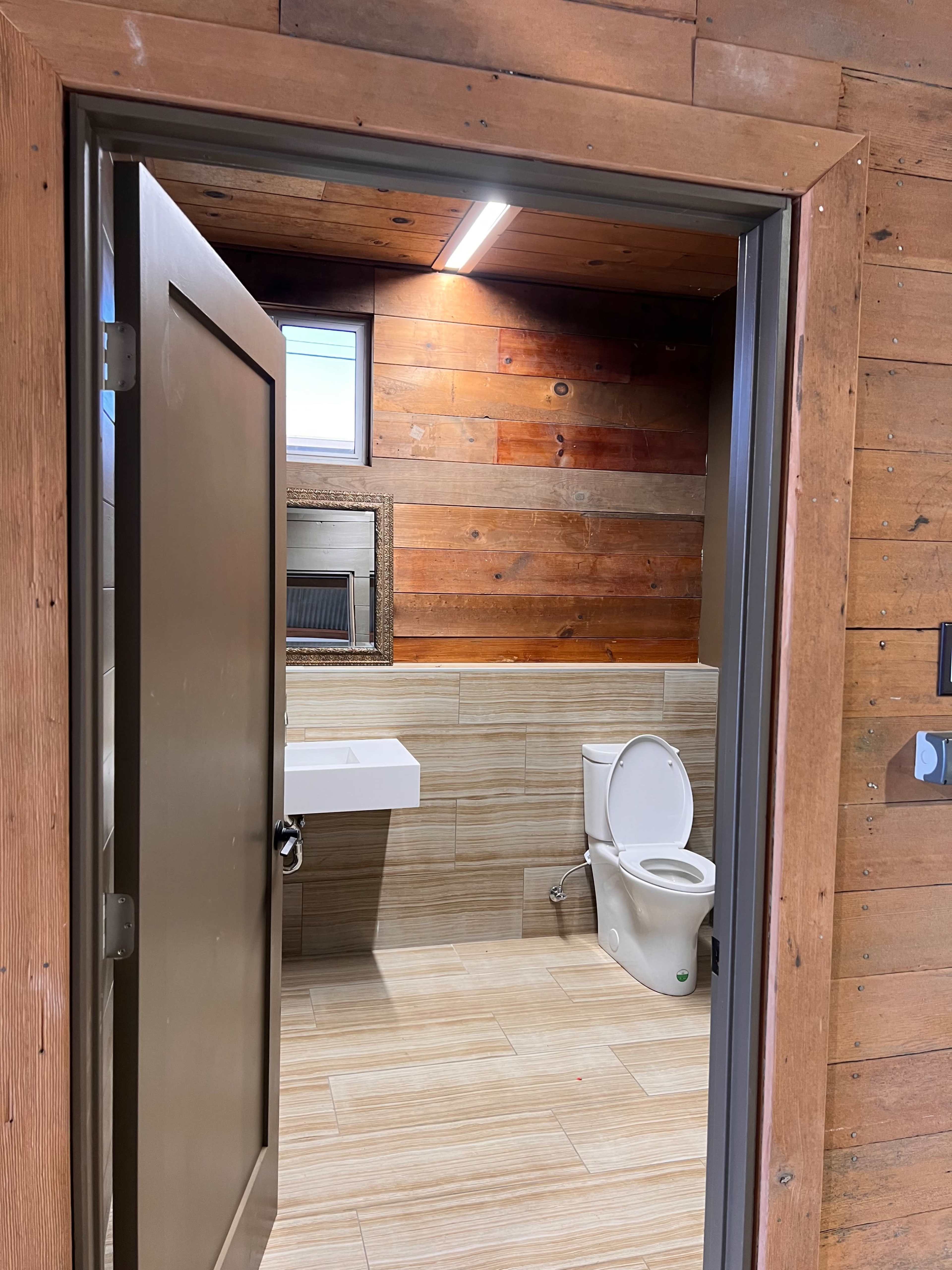 A modern bathroom features a wall-mounted sink, a toilet, and wood-paneled walls, viewed through an open door.