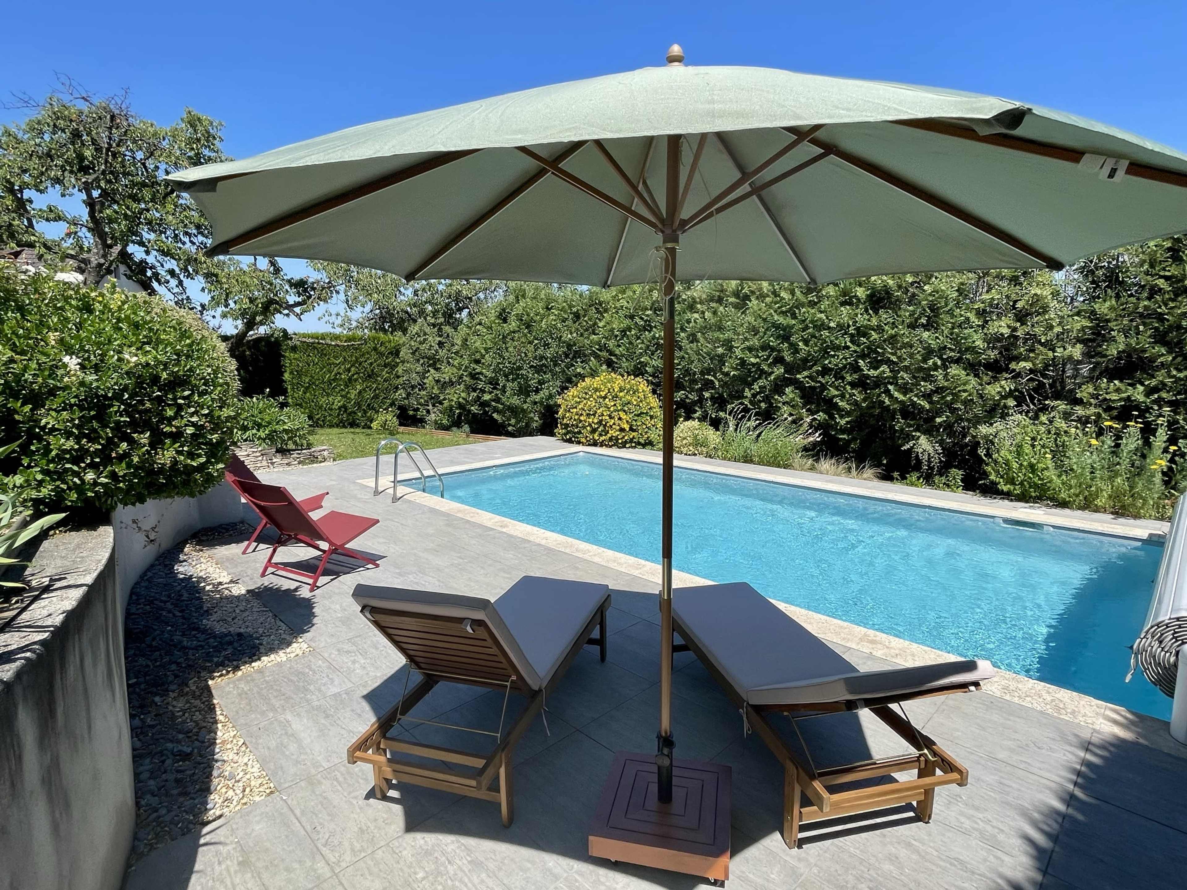 A spacious outdoor pool area features lounge chairs and a large umbrella, surrounded by greenery.