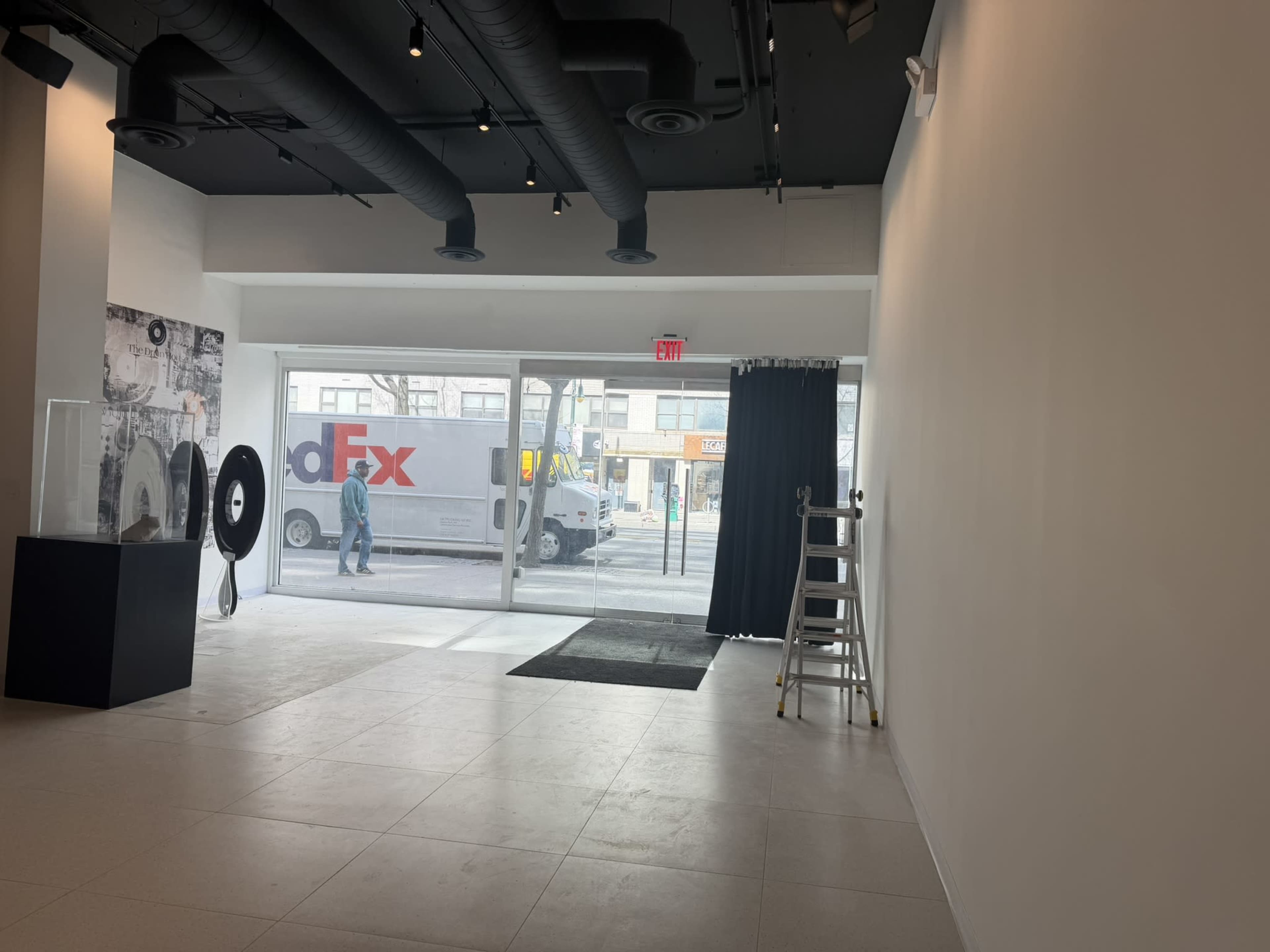 The image shows an empty interior space with a large window displaying a street view, a ladder, and a dark curtain on one side.