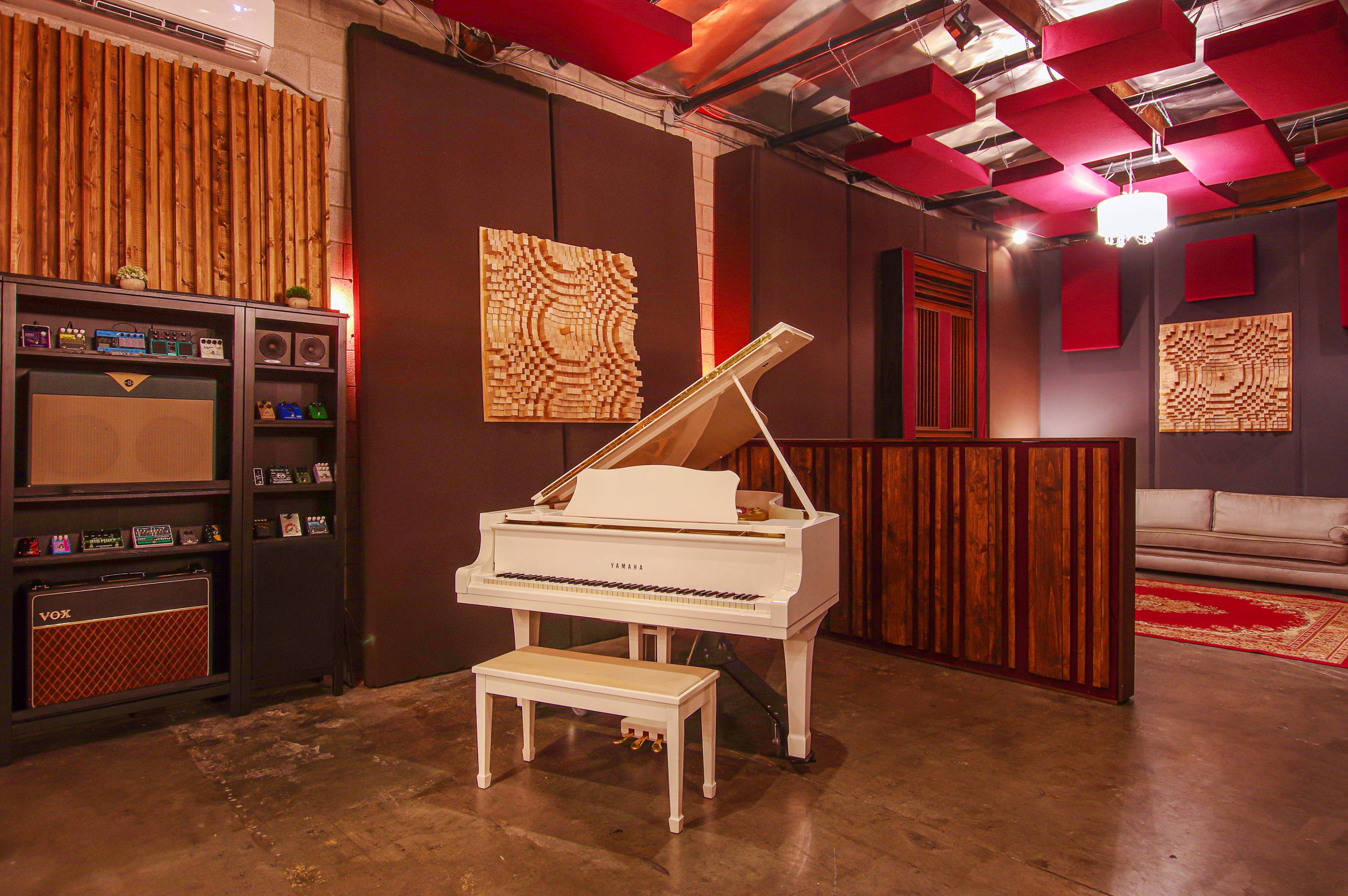 A music studio featuring a white Yamaha piano, a dark shelving unit with audio equipment, and soundproofing panels on the walls.