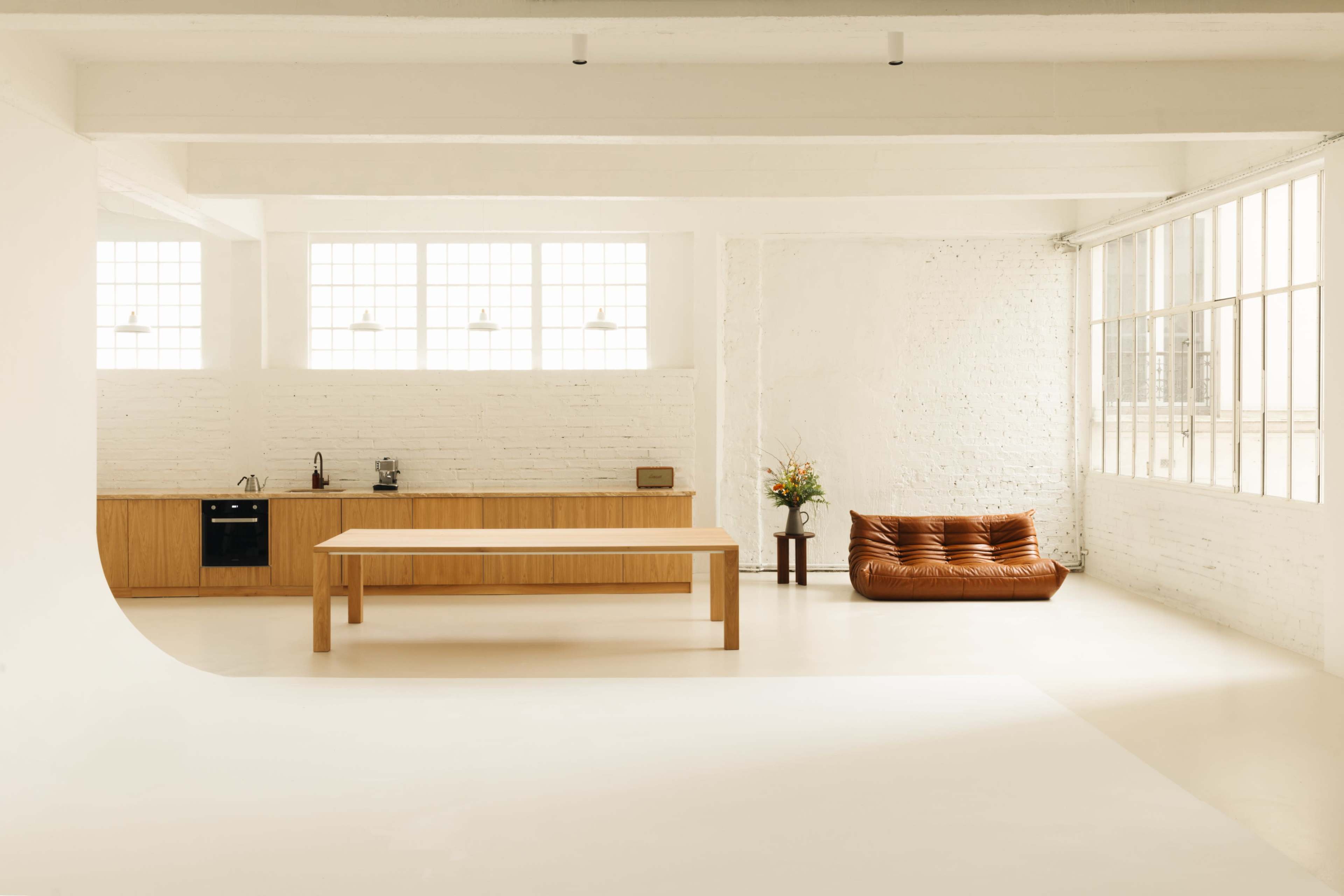 A spacious, minimalist interior with a wooden kitchen, a large dining table, and a brown sofa near a window with natural light.