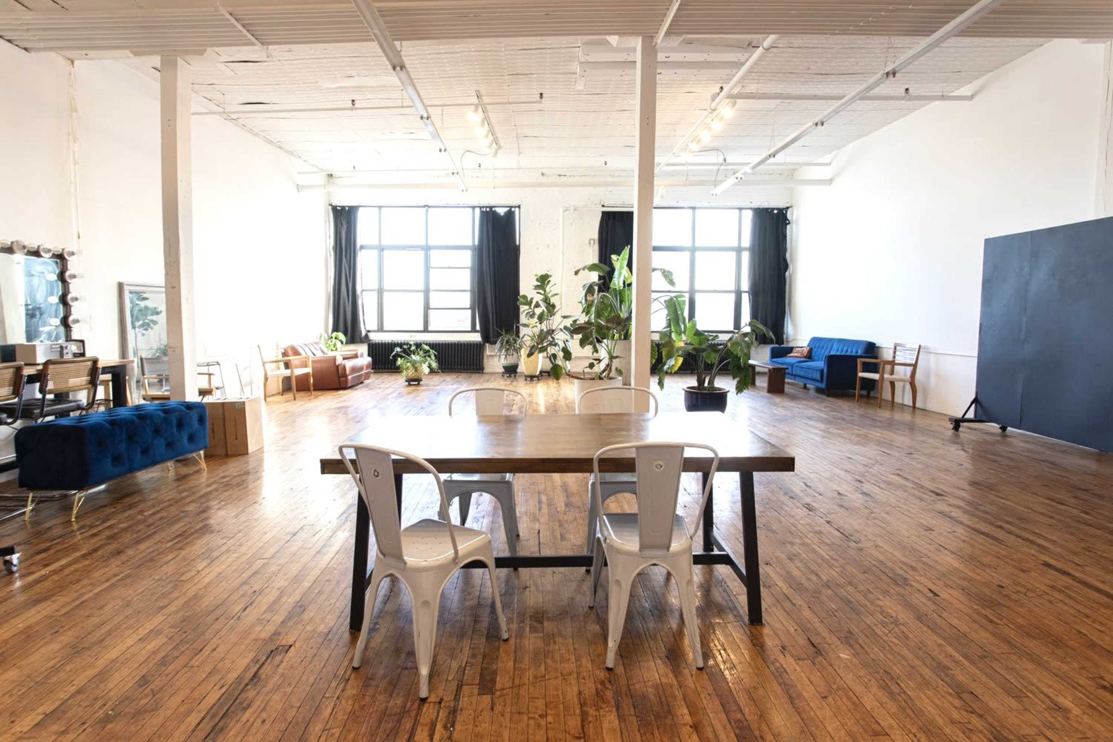 The image shows a spacious, well-lit room with wooden floors, large windows, and various seating arrangements including a long table with metal chairs and several plants throughout.