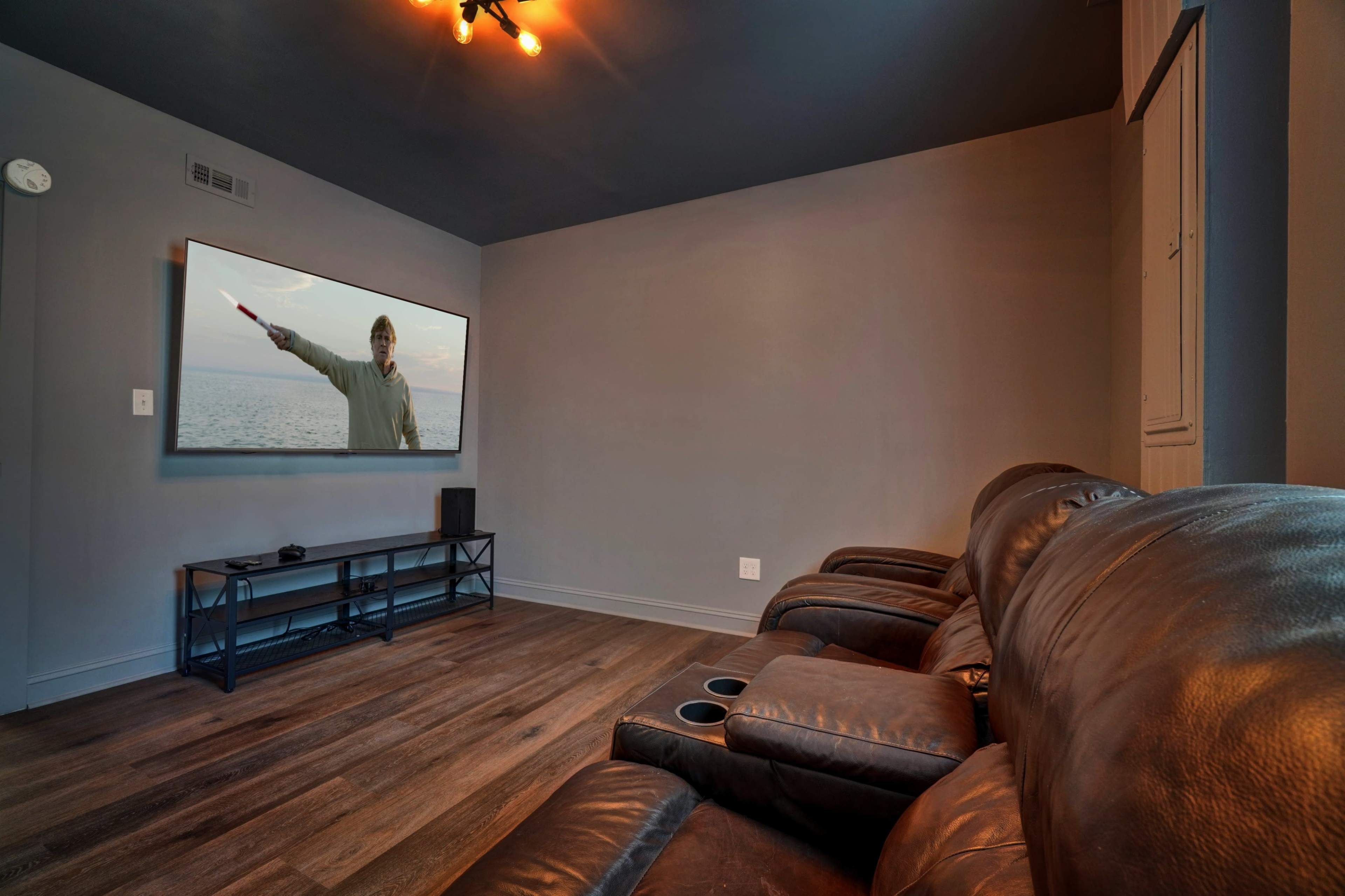 A modern living room features dark walls, a large television showing a scene with a person gesturing, and leather recliners arranged for viewing.