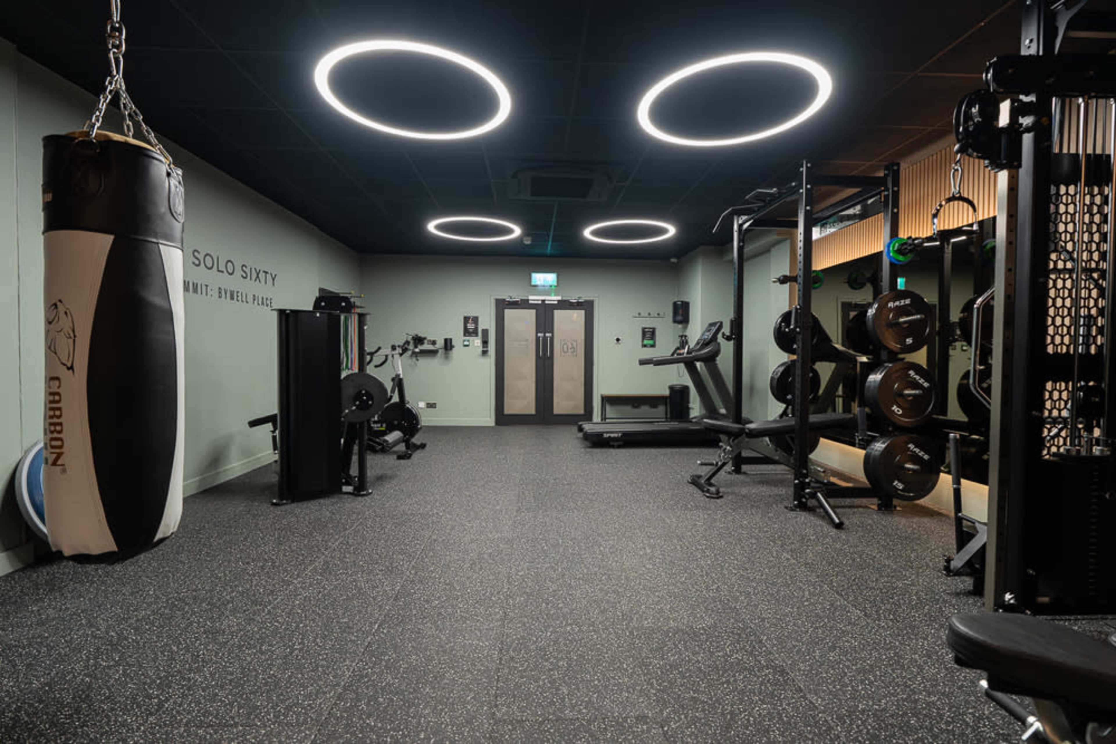 The image shows a modern gym interior featuring exercise equipment, a punching bag, and various weights, all under circular ceiling lights.